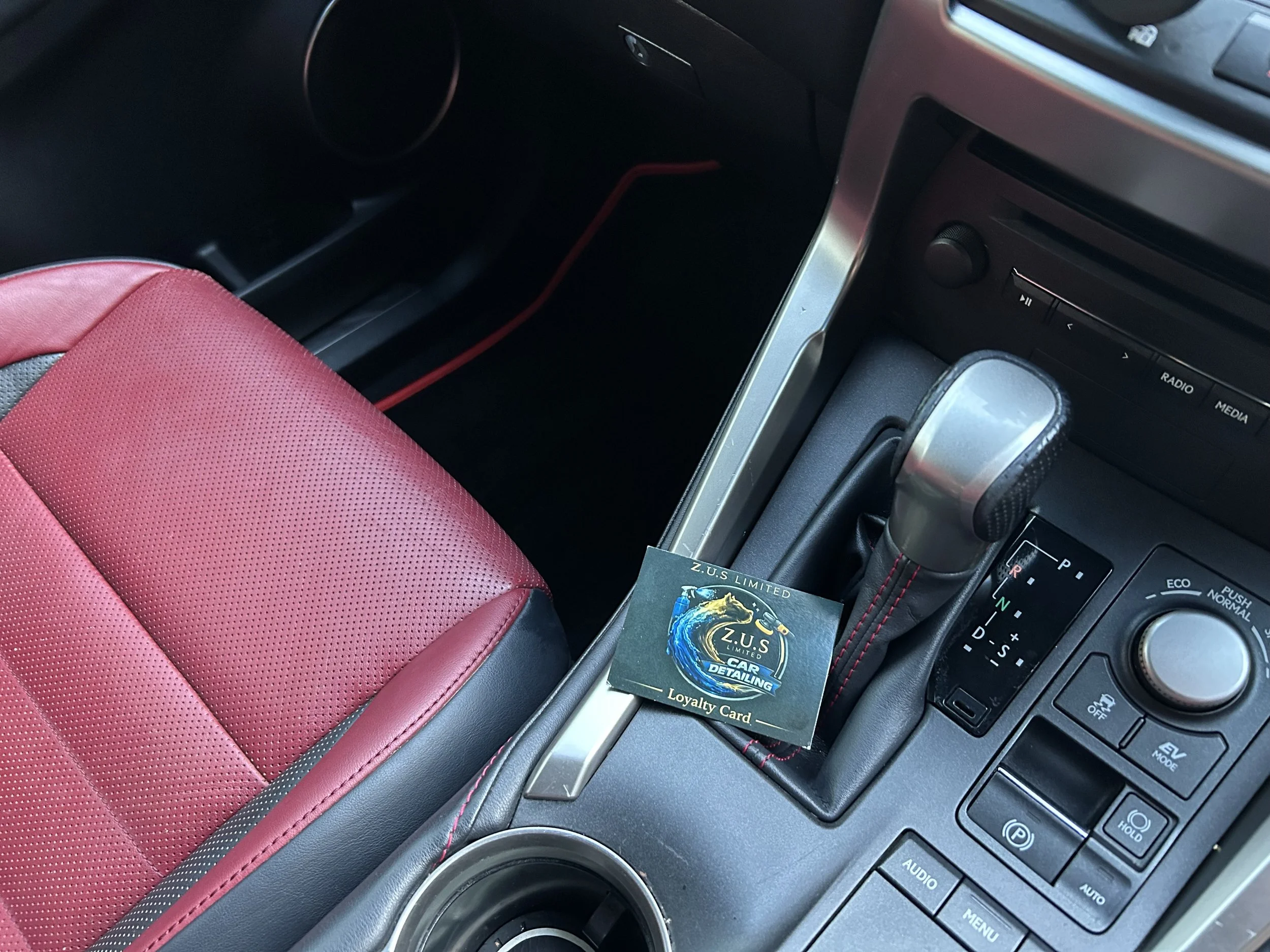 Inside a car, a red and black leather seat, gear shift, and a loyalty card for Z.U.S Limited Car Detailing are visible.