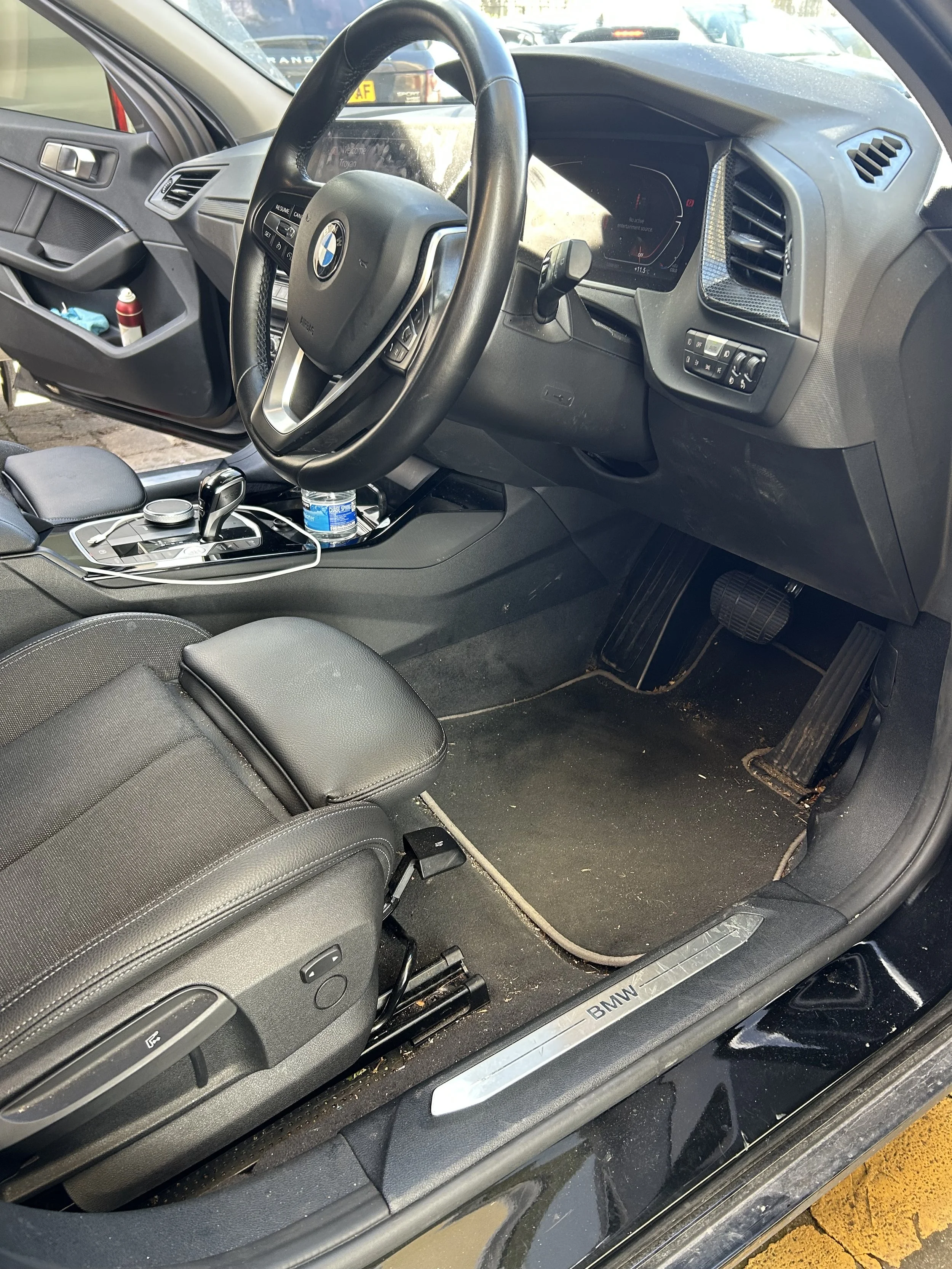 Interior of a BMW car showing the driver's seat, steering wheel, dashboard, center console, and floor area with some dirt and debris.