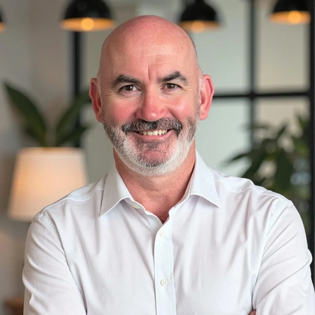A middle-aged man with a bald head, gray beard, and mustache smiling. He is wearing a white dress shirt and standing in a modern office or home setting with plants, a lamp, and black pendant lights in the background.