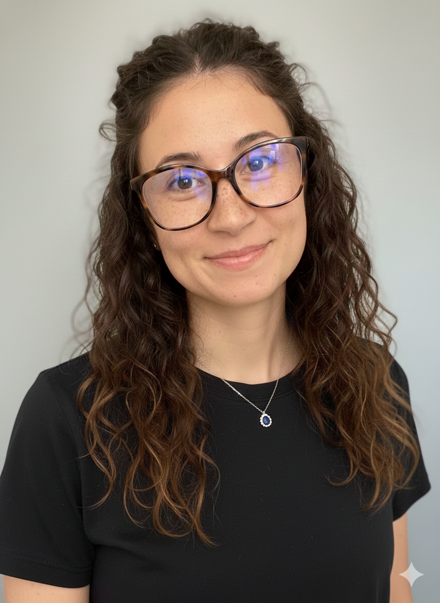 A woman with long, curly brown hair wearing glasses, a black top, and a silver necklace with a circular blue pendant, standing against a plain light-colored background.