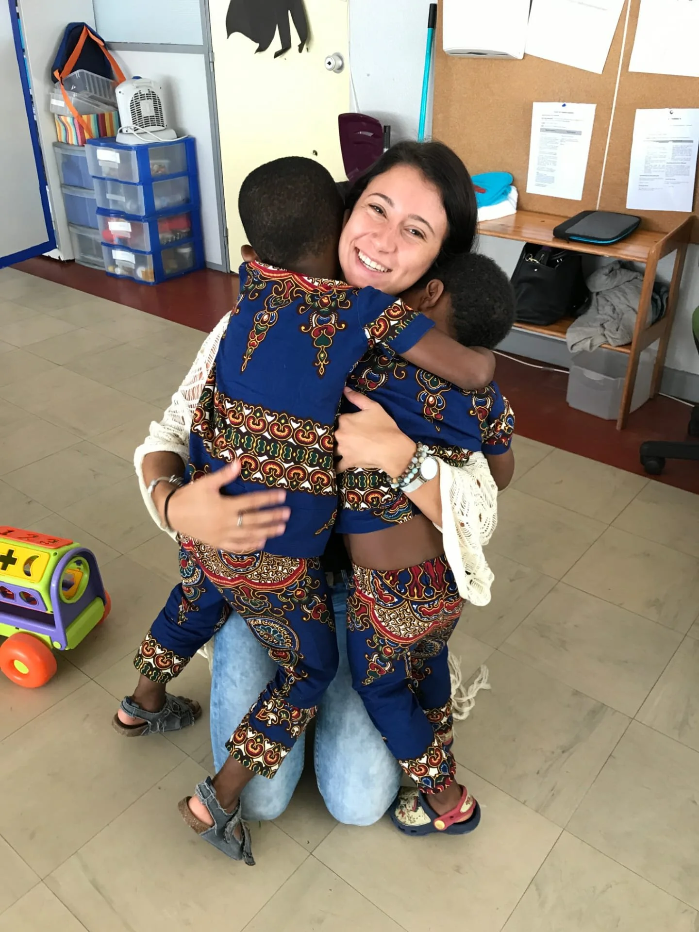 A woman and two children in matching blue and gold patterned clothing hugging indoors, with the woman smiling.