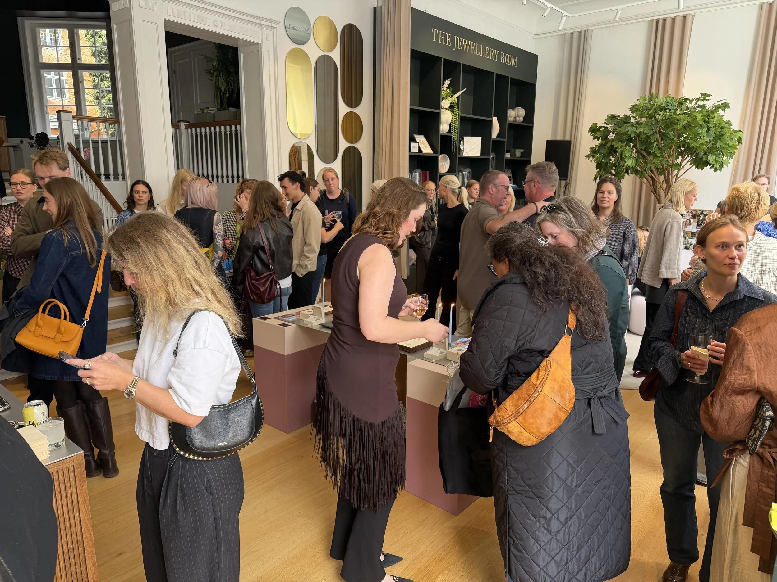 Mennesker i en samling indendørs ved en udstilling eller social begivenhed, nogle med glas vin, foran en sort hylde med dekorative genstande, væggen har et skilt der siger 'The Jewellery Room', store vinduer og grøn plante i baggrunden.