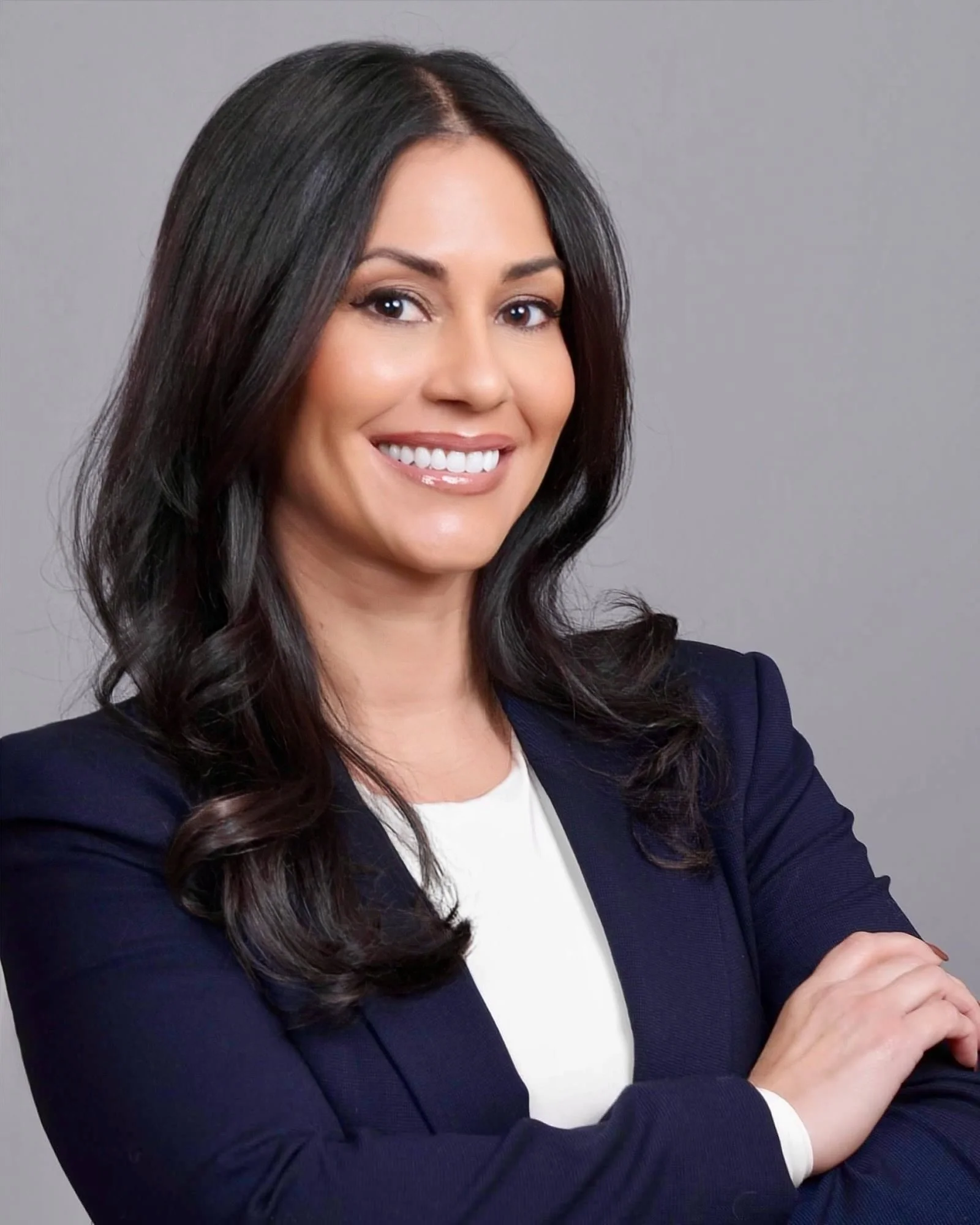 A woman with long dark hair, smiling, wearing a navy blazer over a white shirt, arms crossed, against a gray background.