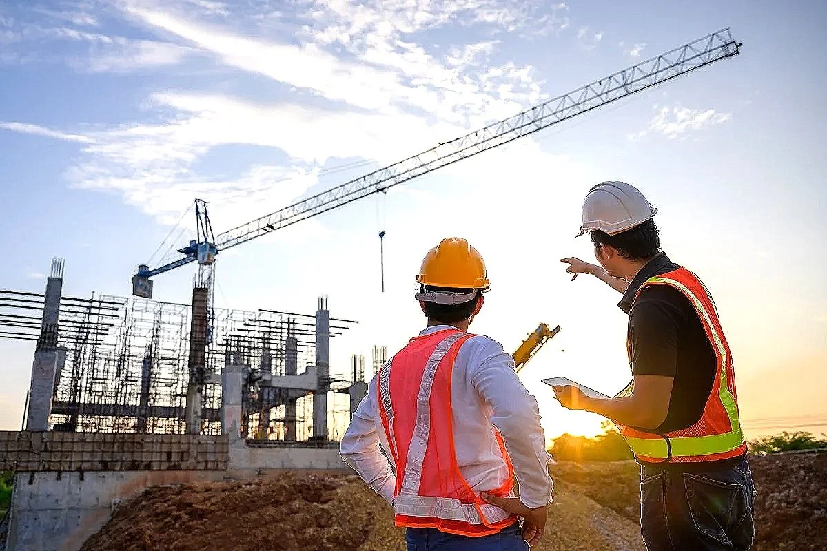 Deux ouvriers de construction portant des gilets de sécurité et des casques discutent sur un site de construction avec une grue en arrière-plan au coucher du soleil.