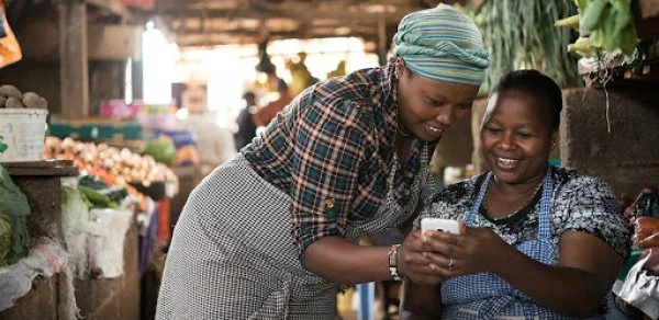 Deux femmes rient en regardant un téléphone dans un marché local.