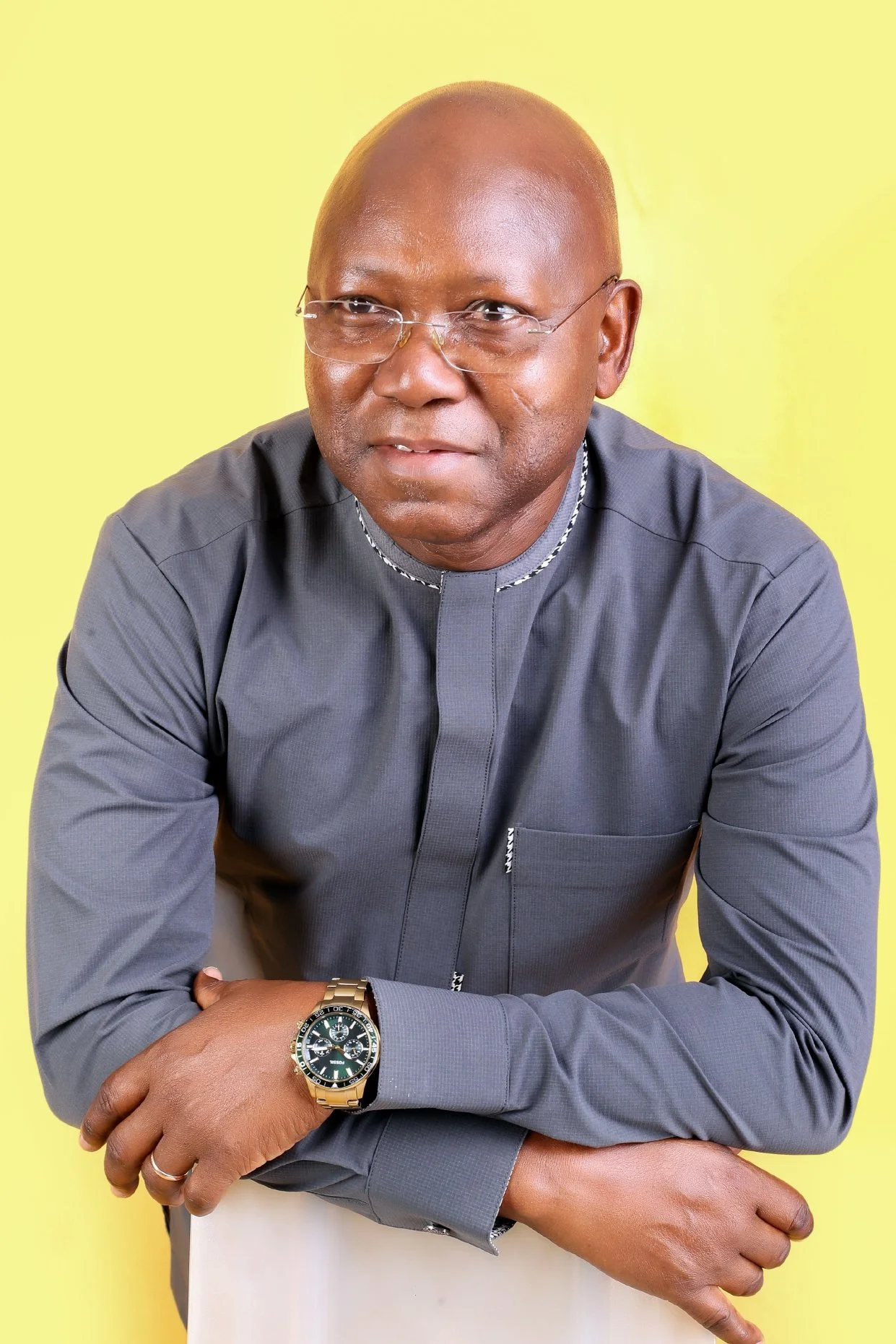 Un homme africain souriant portant des lunettes et une montre en or, habillé en chemise gris foncé, posé contre un fond jaune