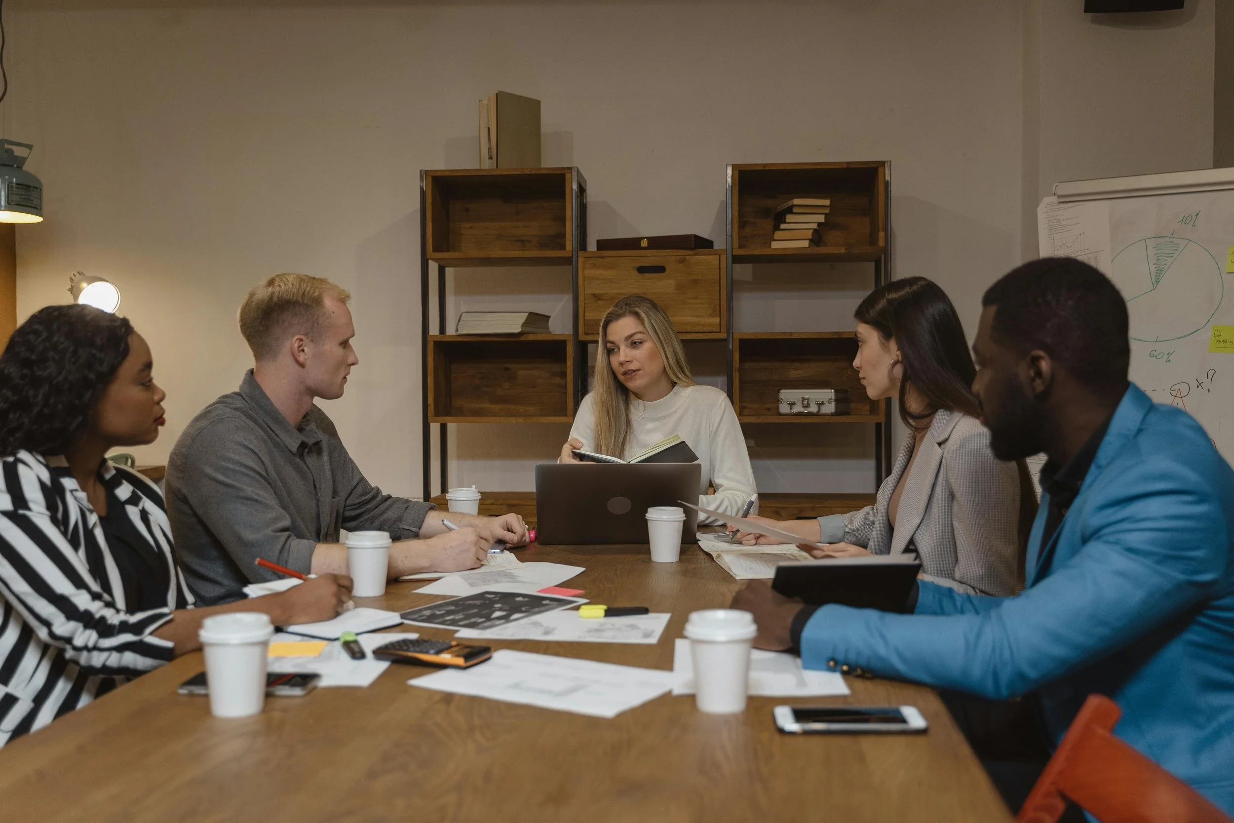 Groupe de personnes en réunion de travail autour d'une table, discutant avec documents, ordinateurs et tasses à café