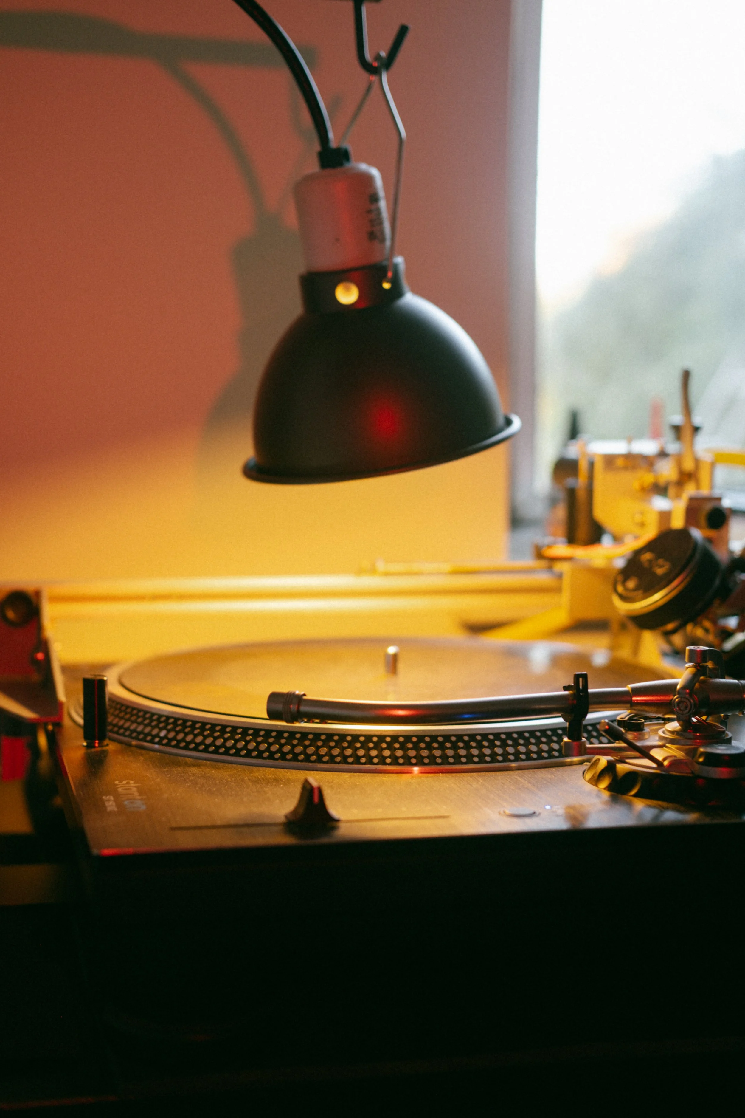 A black turntable with a tonearm on a wooden surface illuminated by a black hanging lamp, with a window in the background showing an outdoor scene.