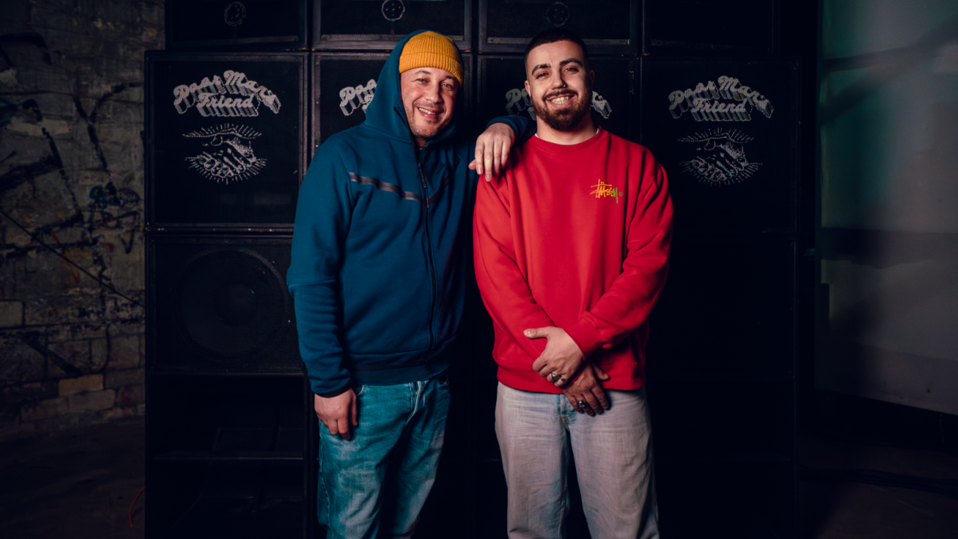 Two men smiling, standing close together in front of a black speaker system, one wearing a blue jacket and yellow beanie, the other wearing a red sweatshirt.
