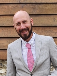 Man with a beard and bald head smiling, wearing a gray suit, white shirt, and pink tie standing outdoors against a wooden fence.