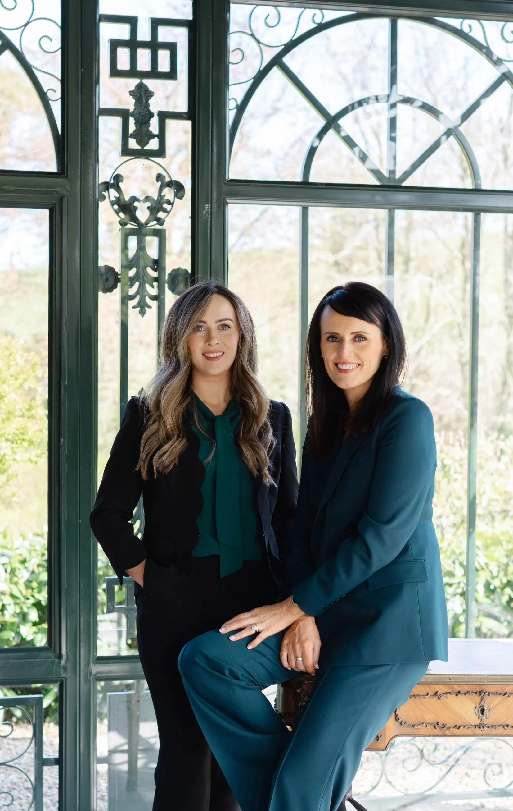 Two women posing together indoors near a large window with decorative ironwork, with outdoor trees in the background. One woman has long wavy blonde hair and the other has short black hair. They are smiling and dressed in professional attire.
