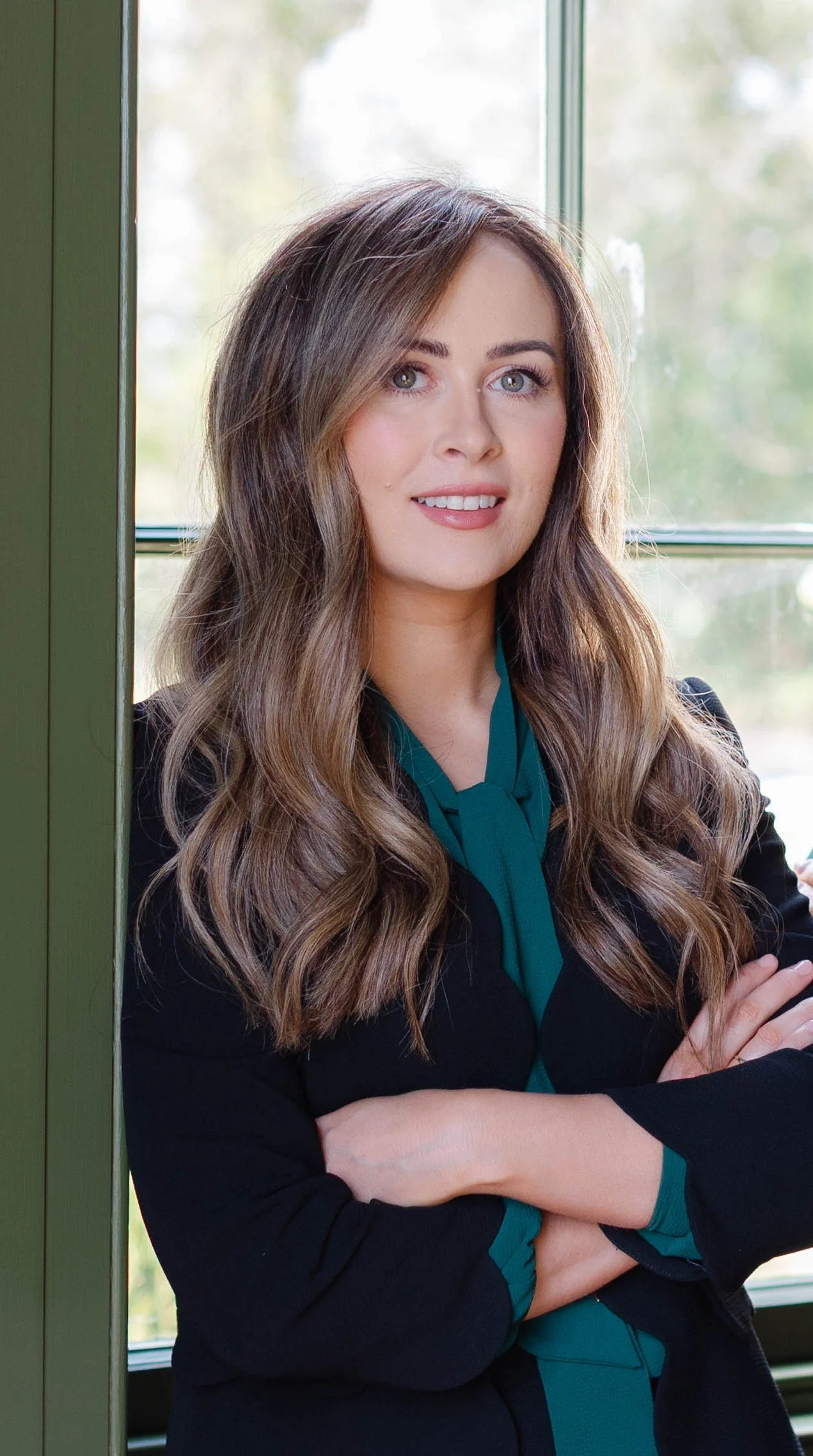 A woman with long wavy brown hair and blue eyes standing near a window with trees outside. She is smiling slightly and has her arms crossed. She is wearing a teal shirt under a black blazer.