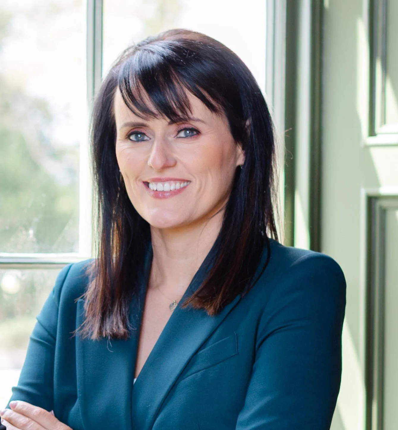 A woman with dark brown hair and blue eyes smiling, wearing a dark blazer, sitting near a window with greenery outside.