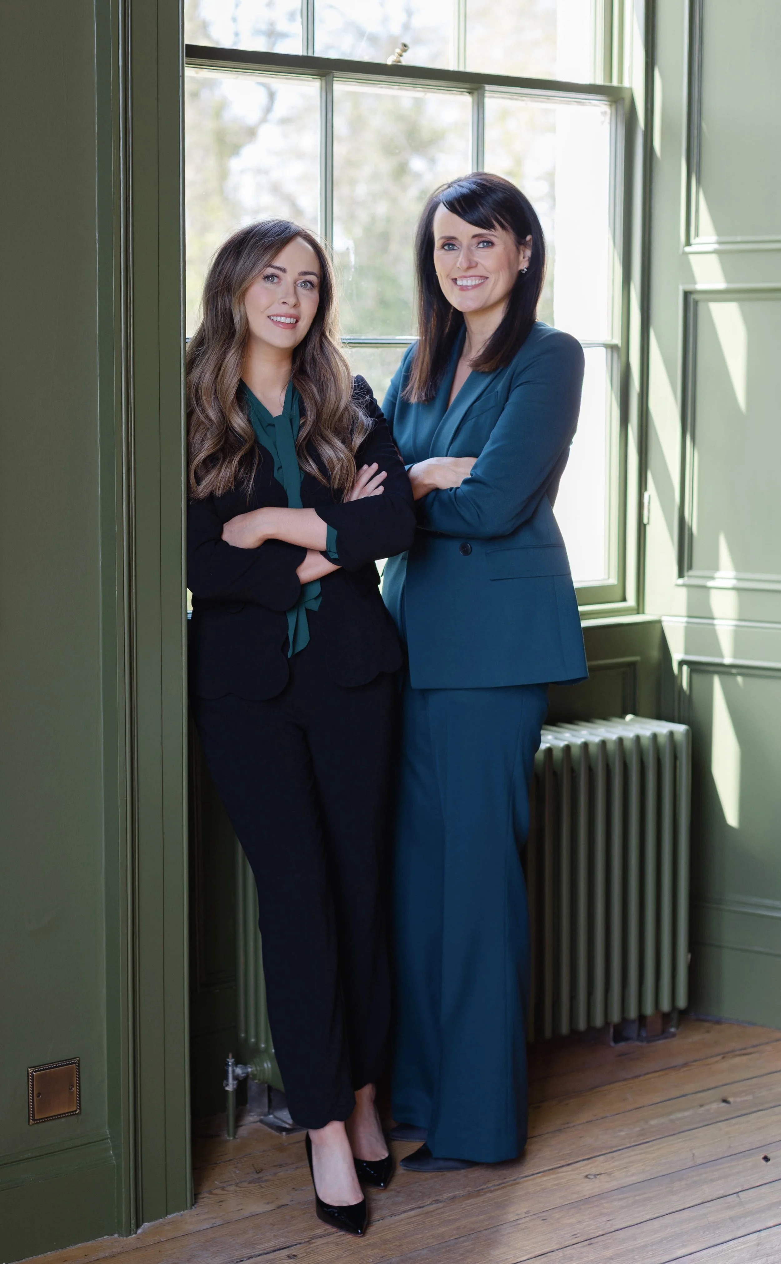 Two women standing next to each other on a wooden floor in front of a green wall and window, smiling at the camera, arms crossed, dressed in formal business attire.