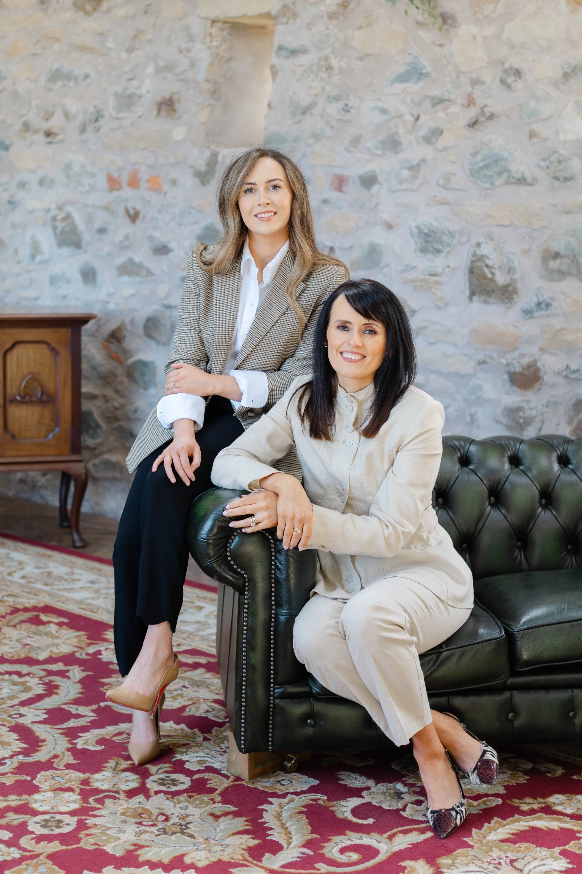 Two women in formal attire sitting on a black leather tufted sofa in a room with stone walls and a wooden side table. One woman, with brown hair and a beige suit, is sitting on the sofa arm. The other woman, with black hair and a white suit, is seated on the sofa. Both women are smiling, and the room has a patterned red and beige rug.