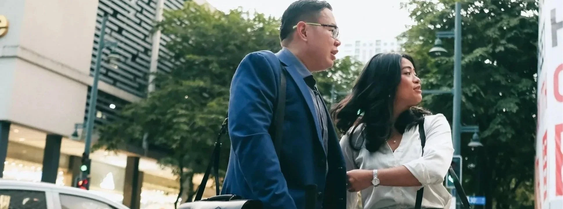 A man and woman walking outdoors in an urban area, with trees and buildings in the background. The man is wearing glasses and a blue blazer, while the woman is dressed in a white blouse and carrying a backpack.