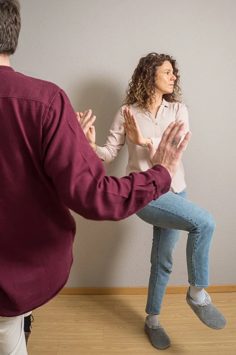 A woman practicing a yoga pose against a plain wall while a man assists or guides her.