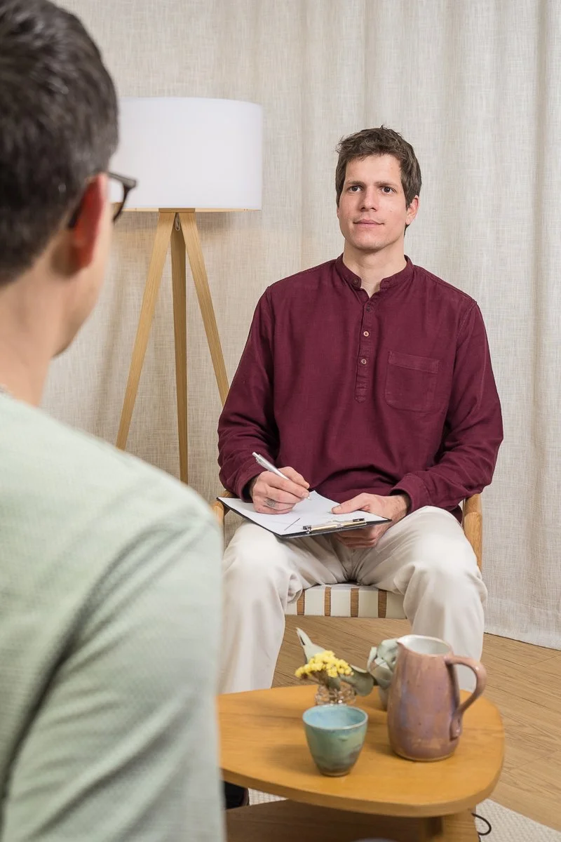 Ein junger Mann in einem weinroten Hemd und hellen Hosen sitzt während einer Therapiesitzung auf einem Stuhl und hält Klemmbrett und Stift in der Hand. Der Raum hat beige Wände, einen kleinen Holztisch mit einem Krug und einer Blume.