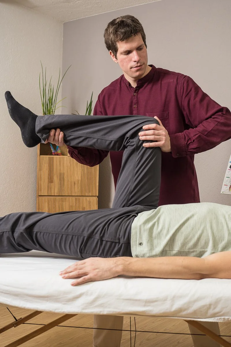 A man in a maroon shirt is holding a person's leg during a physical therapy session in a clinic or medical room.