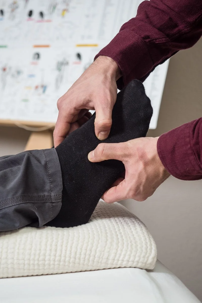 Eine Person hält und betrachtet einen Fus, der von einer schwarzen Socke bedeckt ist. Die Person trägt ein weinrotes Langarmhemd, und der Fuß scheint auf einem strukturierten weißen Kissen zu liegen. Im Hintergrund seht eine Kiensiologie Tafel