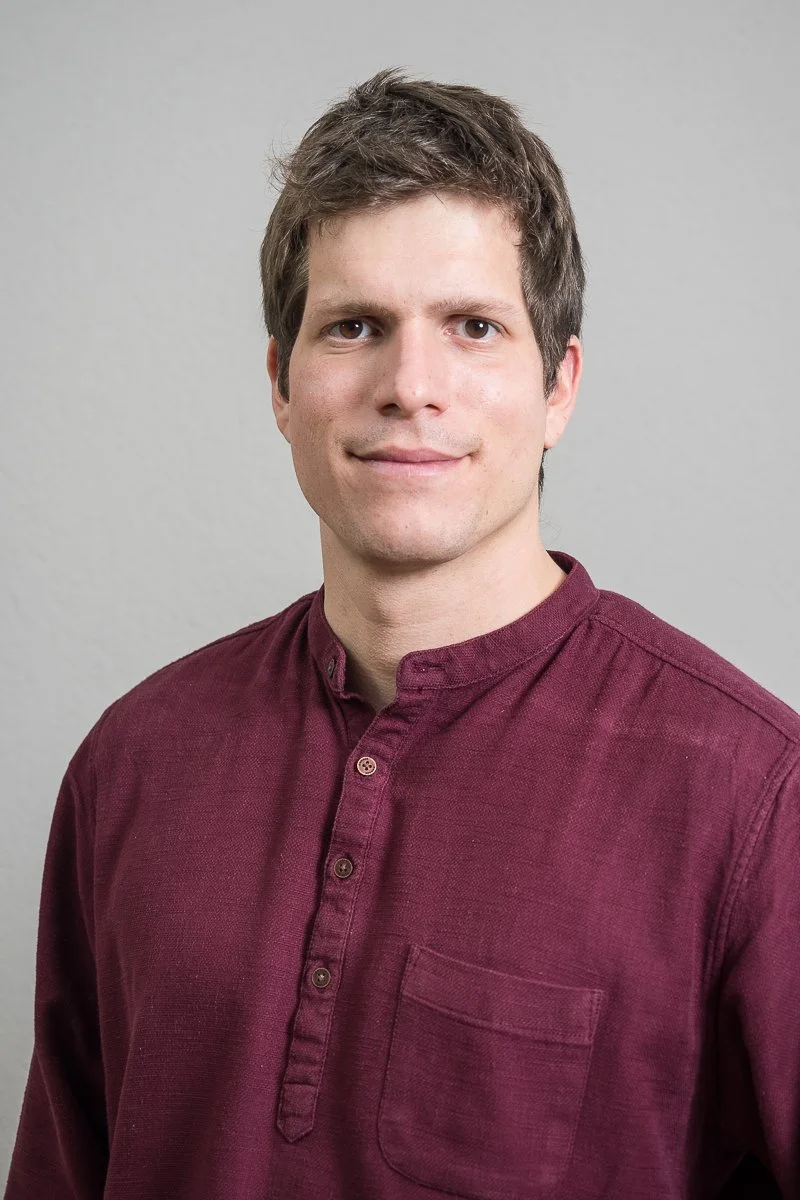 Portrait of a young man with light skin, brown hair, and wearing a maroon button-up shirt, smiling at the camera against a plain gray background.