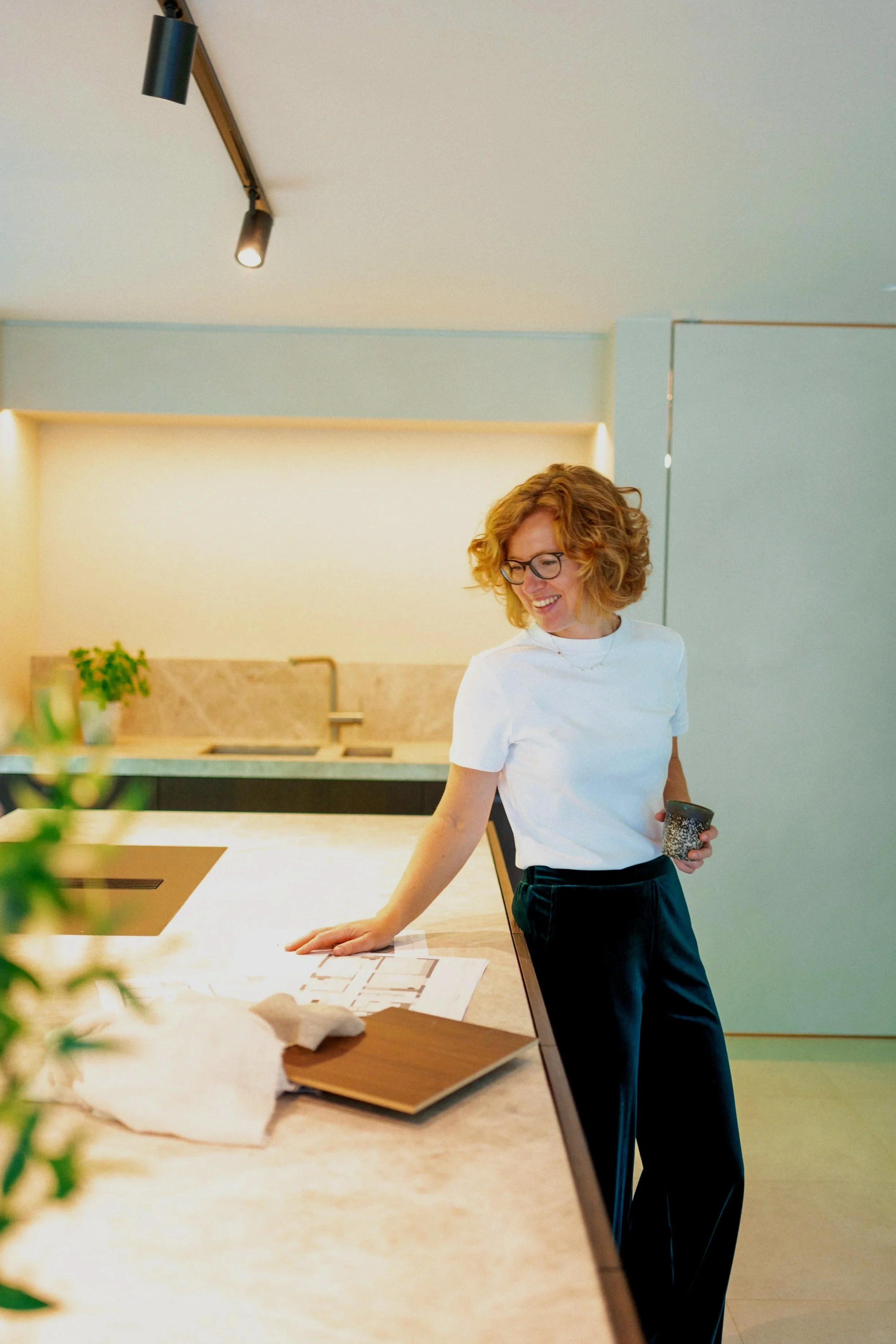 Woman with curly red hair and glasses smiling in a modern kitchen, holding a cup and looking at papers on the kitchen island.