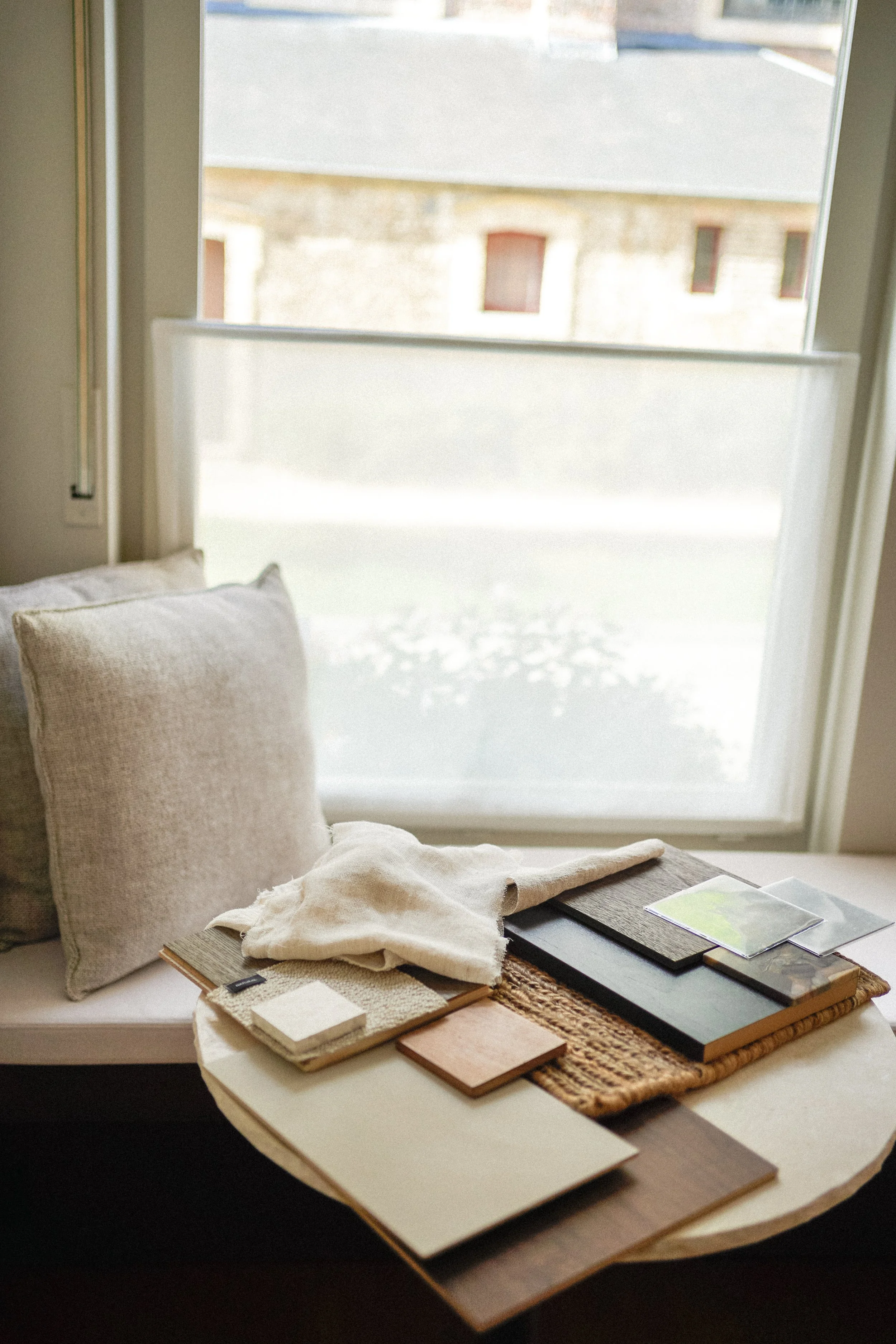 A window seat with beige pillows and a round wooden table displaying various fabric samples, color swatches, and design materials, with sunlight streaming through a large window.