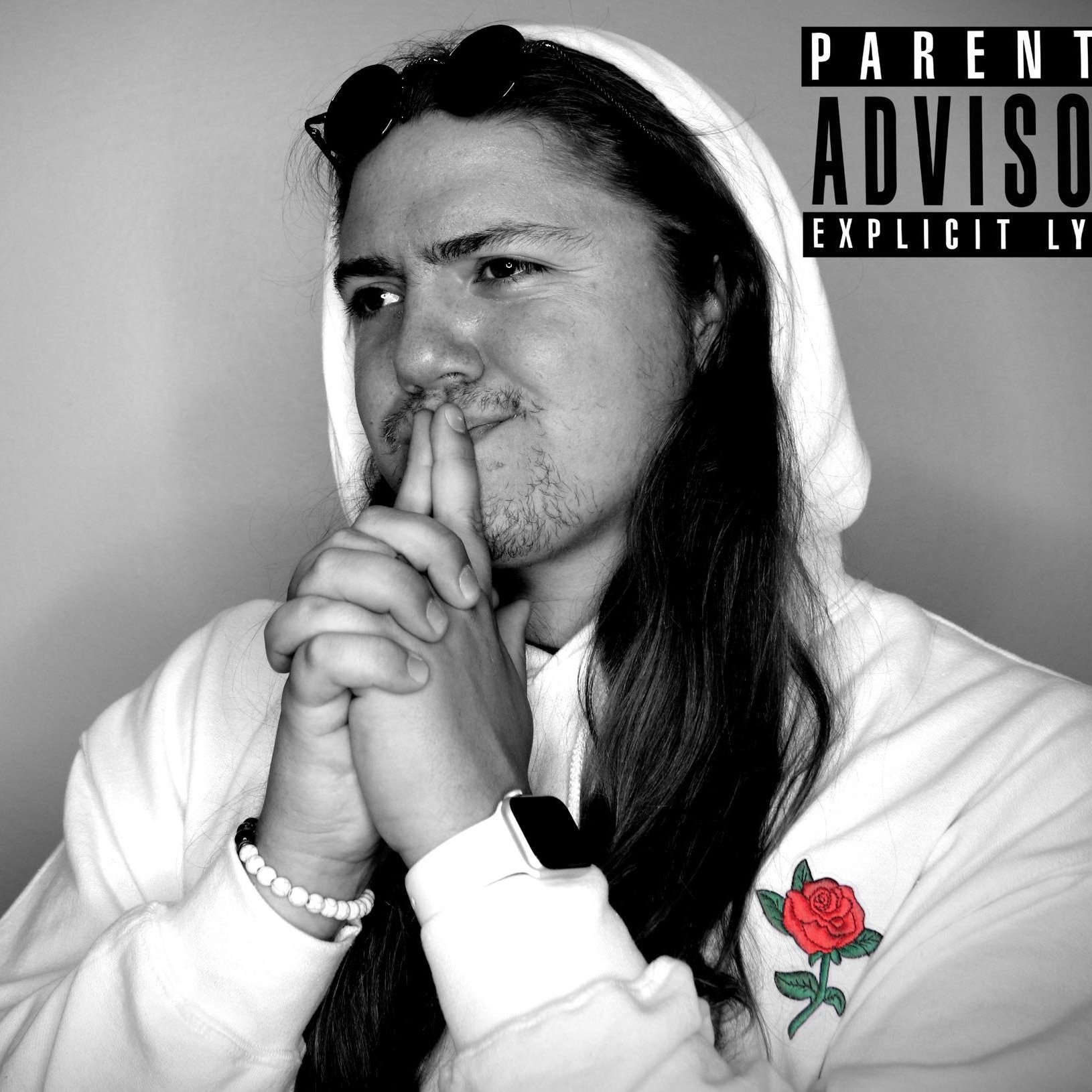 Black and white photo of a man with long hair, wearing a hoodie with a rose patch on the chest, and a smartwatch on his wrist, holding his hands together near his face in a contemplative pose, with a 'Parental Advisory' warning graphic in the top right corner.