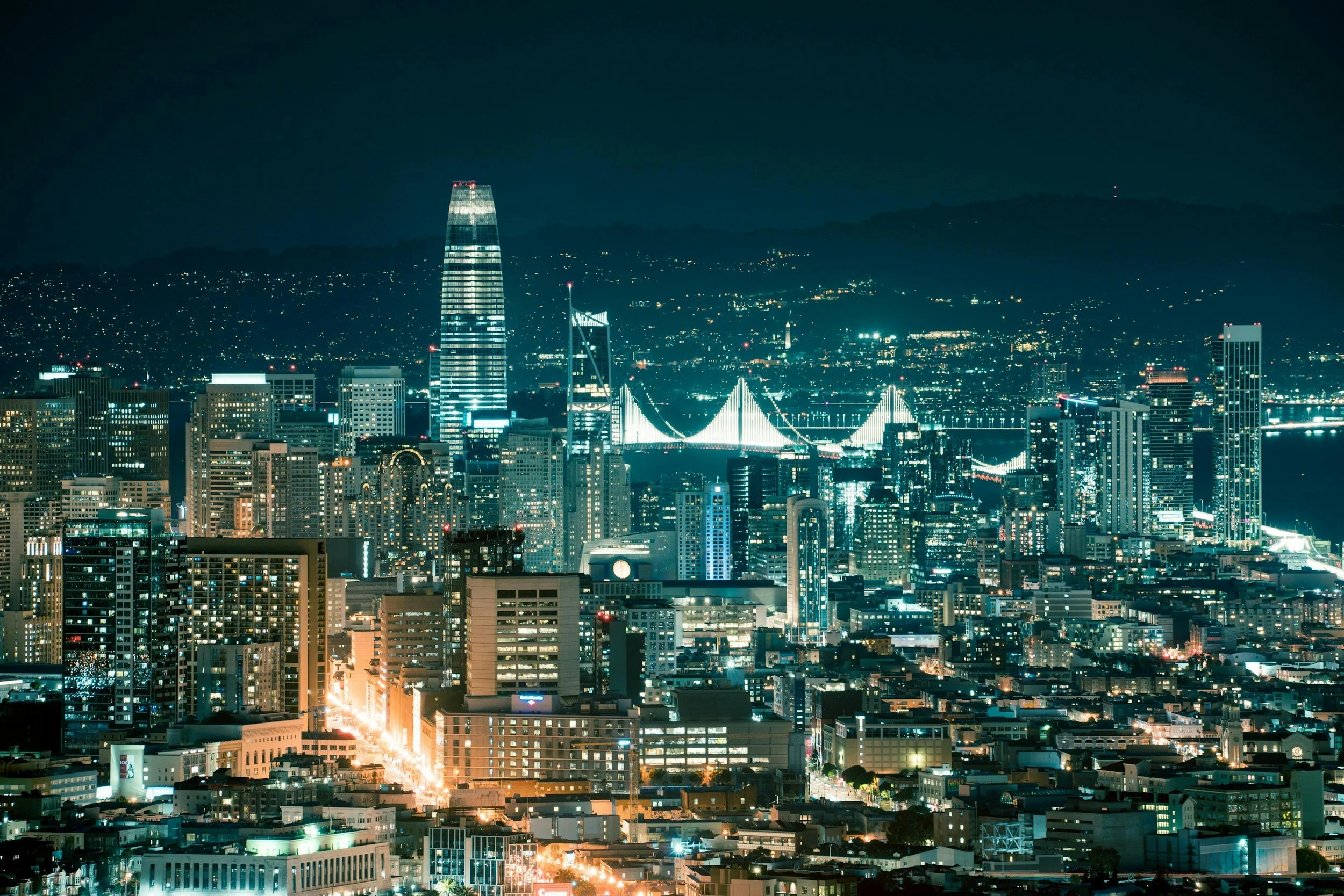 Night view of a city skyline with tall buildings, illuminated bridges, and mountain in the background.