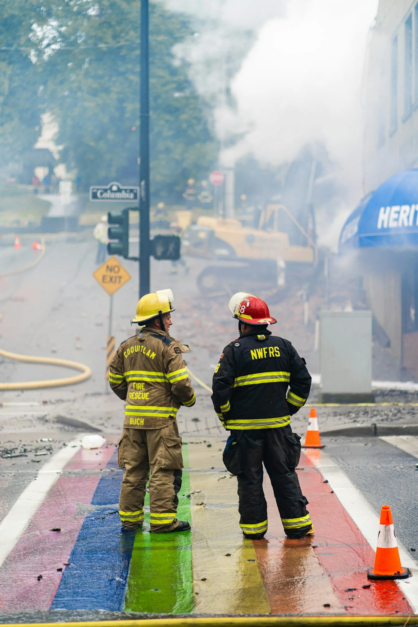 Firefighters stand on a rainbow-colored crosswalk with debris and damage around, smoke in the background, and a fire truck.