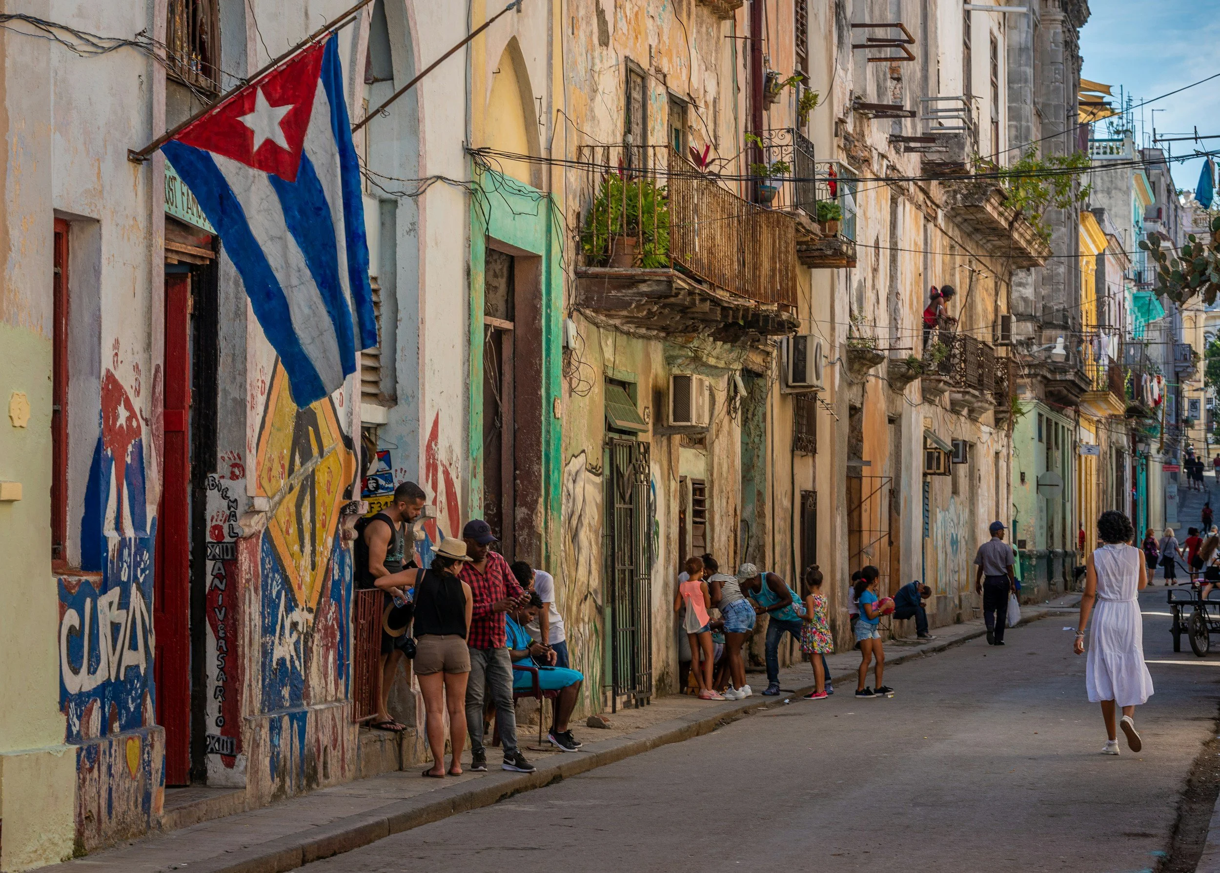 Street scene in Havana, Cuba, with colorful colonial-style buildings, a hanging Cuban flag, and people walking and standing along the sidewalk.