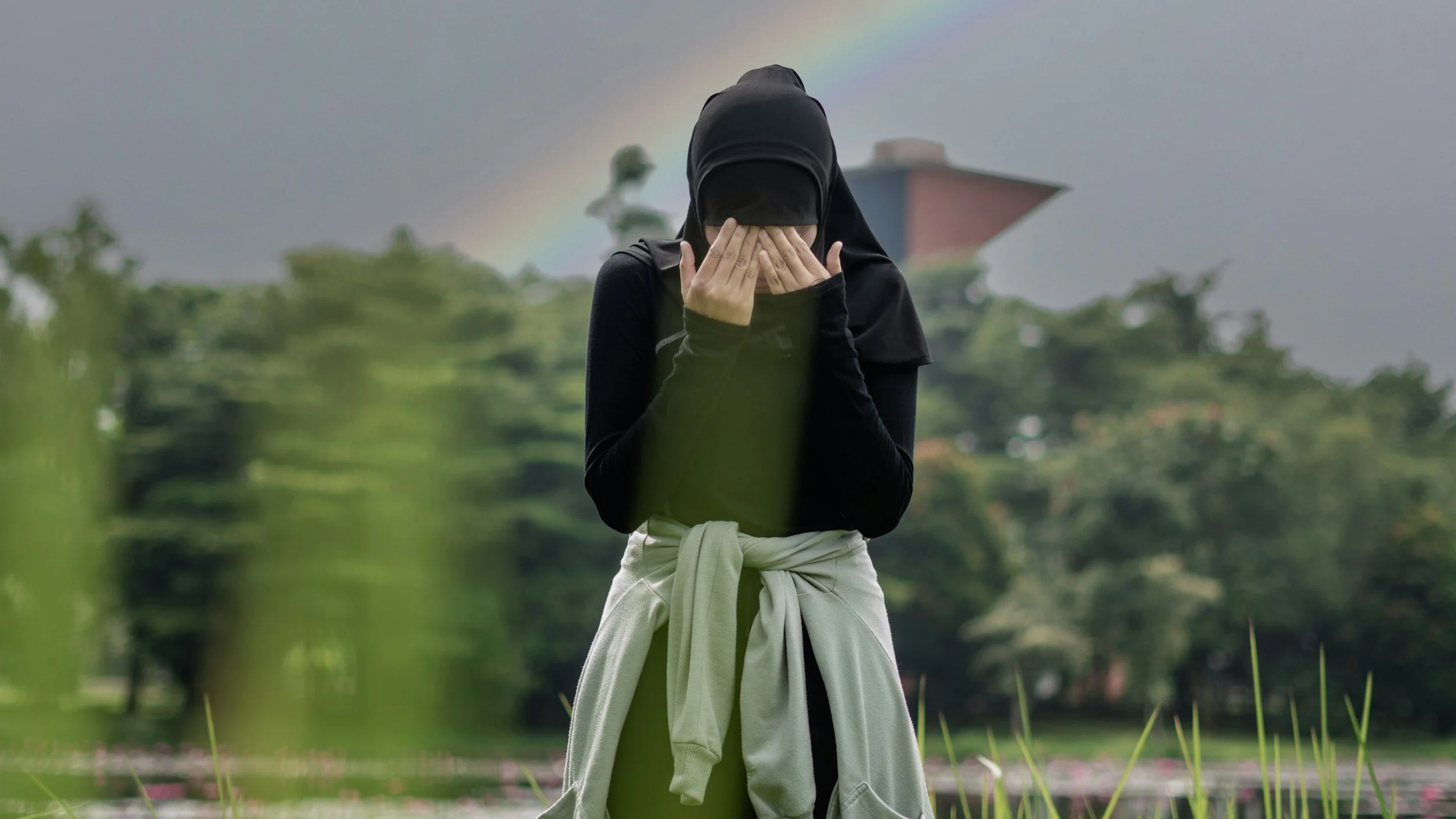 A person wearing a black hoodie and black pants, covering their face with their hands, standing outdoors with a rainbow in the cloudy sky and greenery in the background.