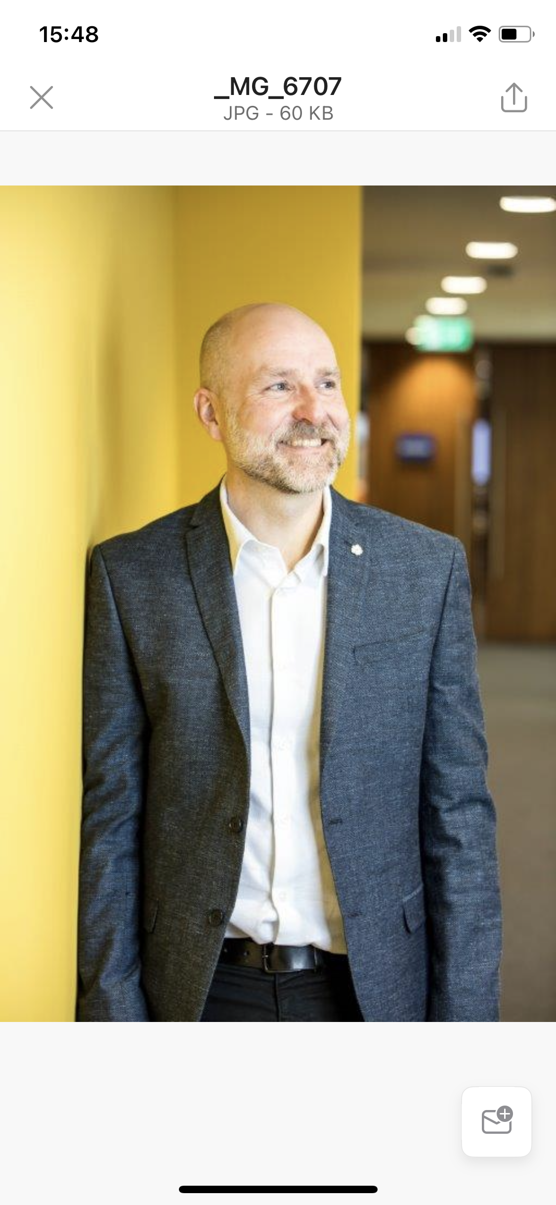 A man with a beard and bald head is smiling while leaning against a yellow wall. He is wearing a dark grey blazer, a white shirt, and black pants, in an indoor setting with blurred background lights.