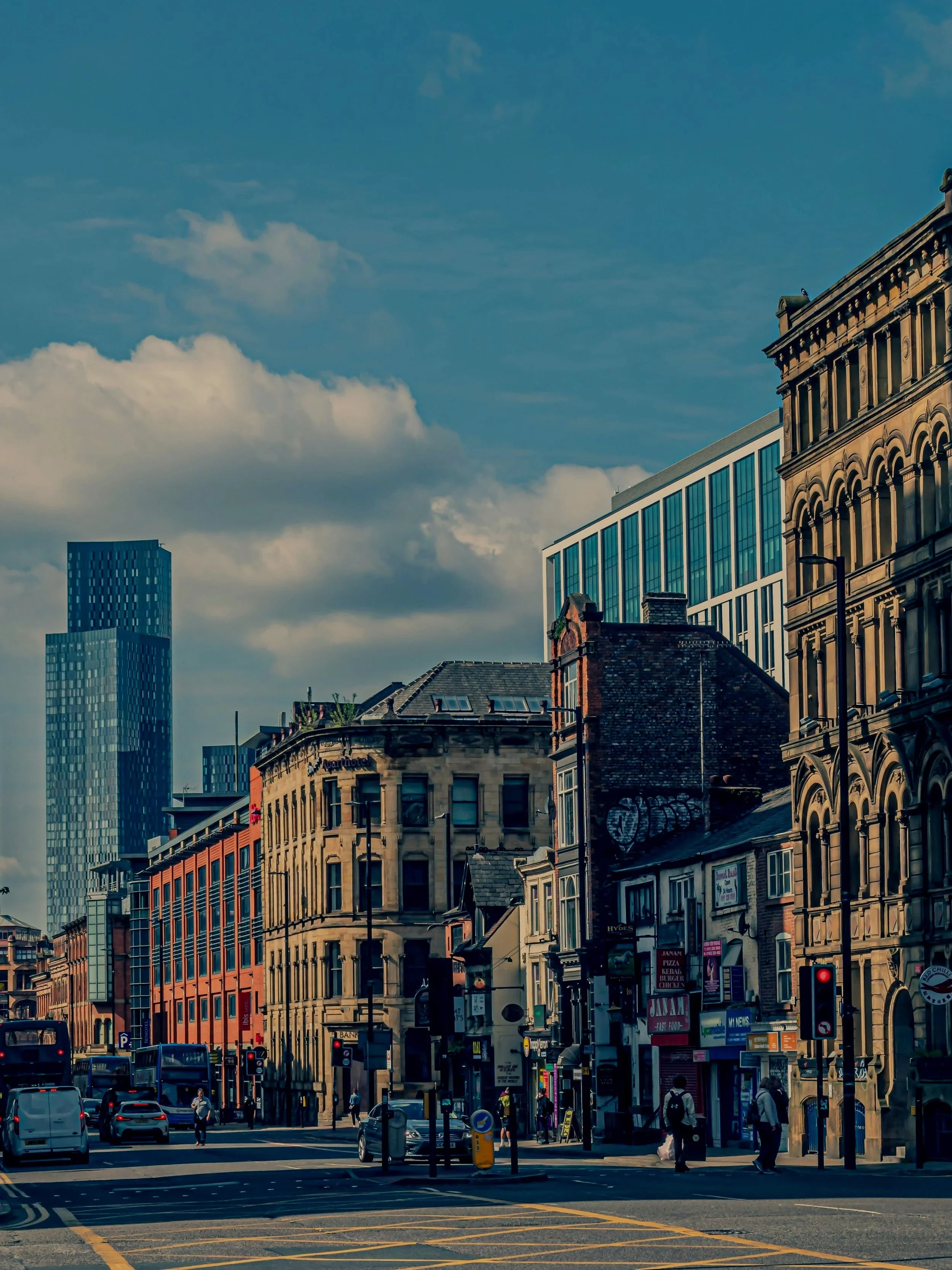City street scene with historic and modern buildings, cars, buses, pedestrians, and traffic lights under a partly cloudy sky.