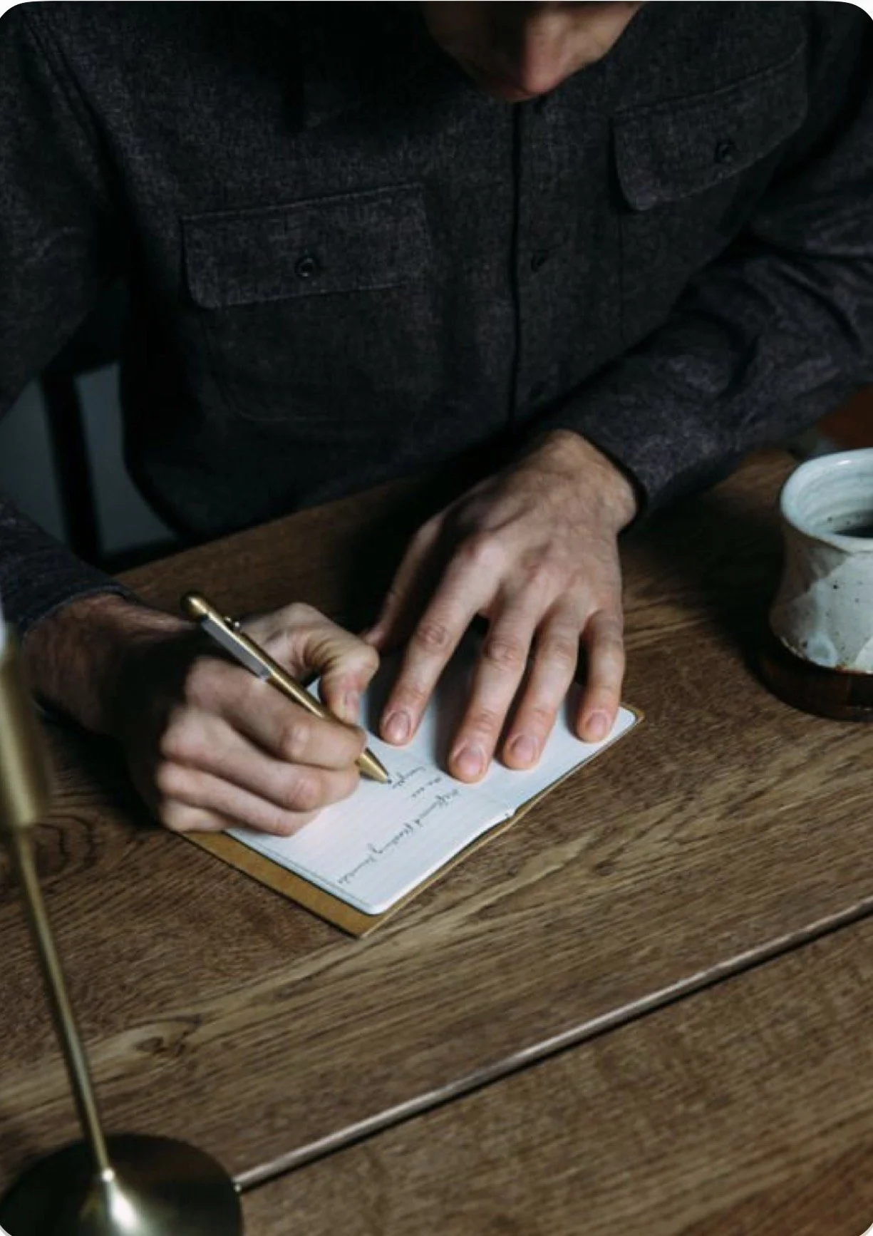 A person writing notes in a notebook with a gold pen, sitting at a wooden table with a cup of dark coffee nearby.