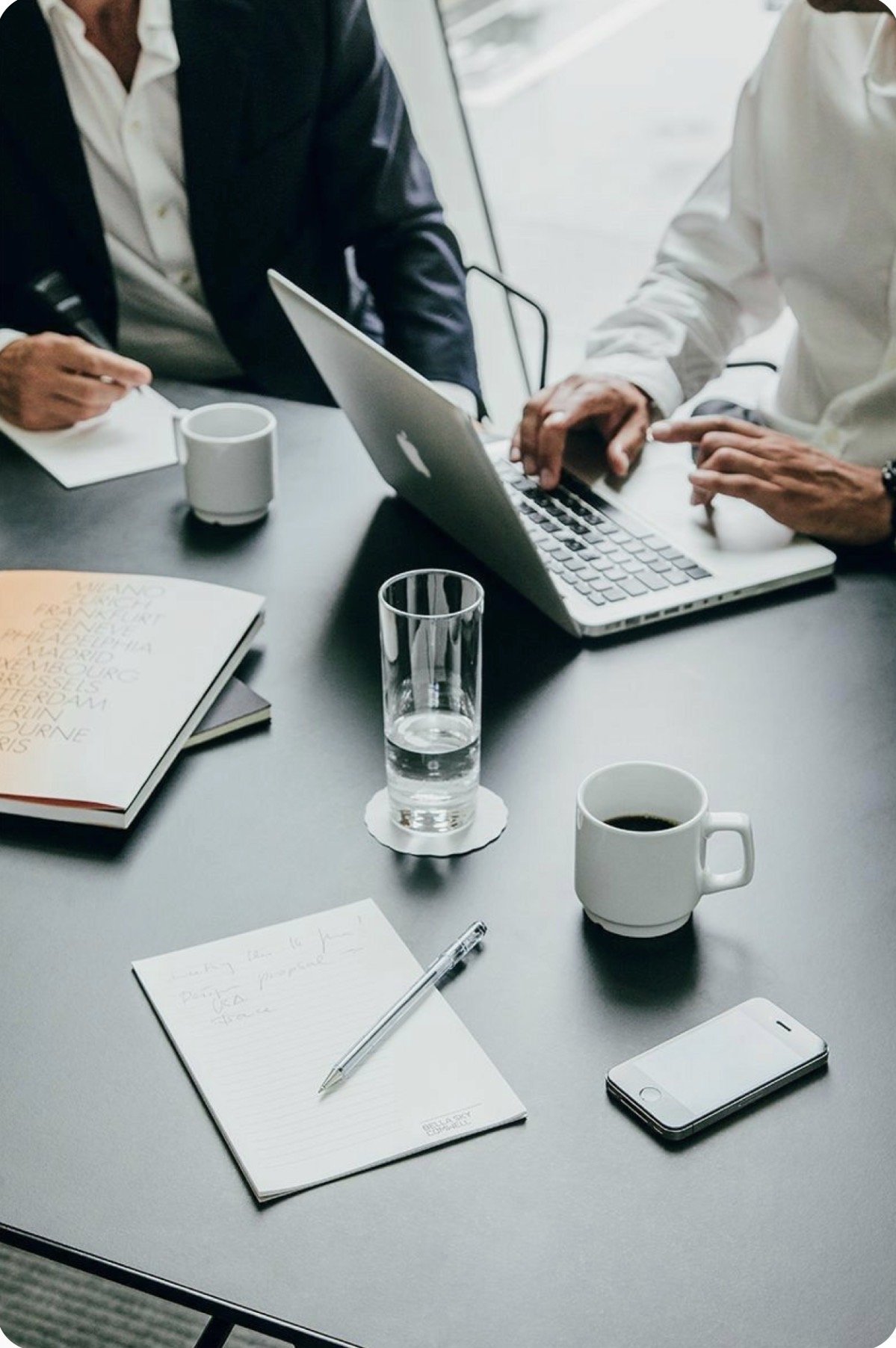 Two people work together at a conference table with a laptop, notebooks, coffee mugs, a glass of water, a pen, and a smartphone.