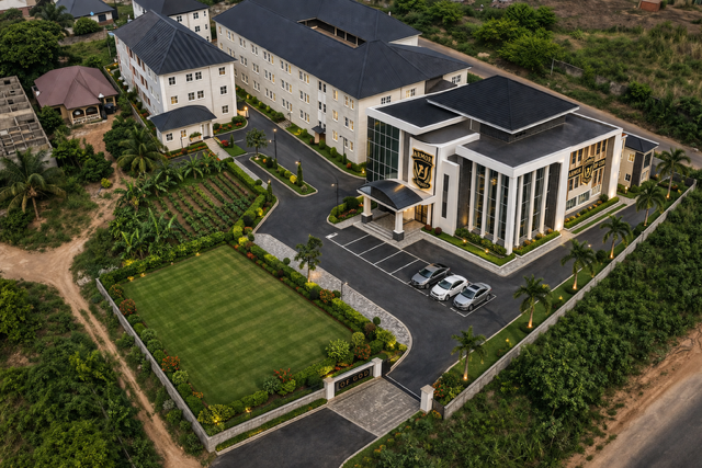 A large modern building with an estate and parking lot, surrounded by landscaped greenery, trees, and a sports field.