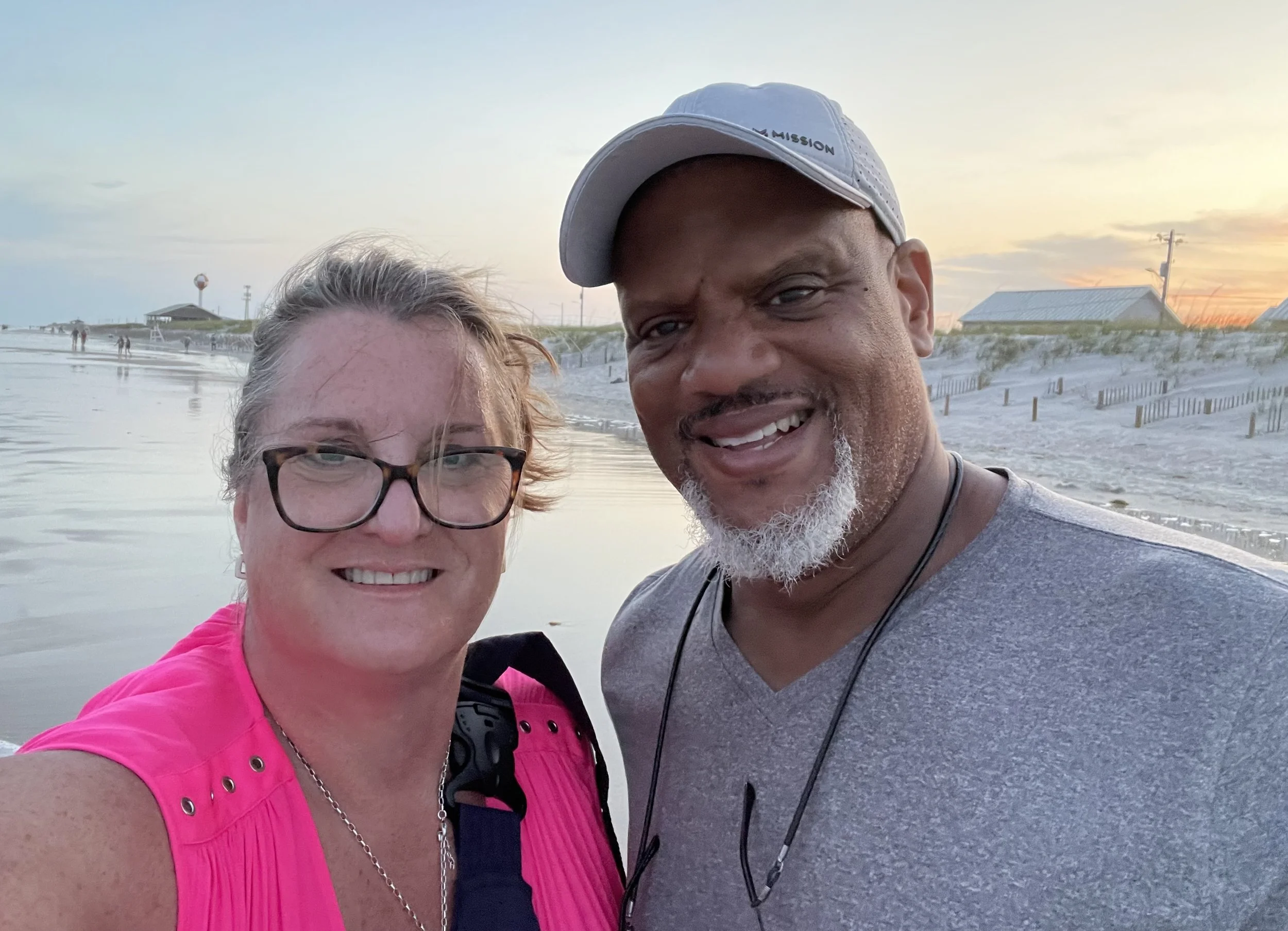 A smiling woman with glasses and a pink shirt standing next to a smiling man with a gray shirt and a khaki cap at the beach during sunset.