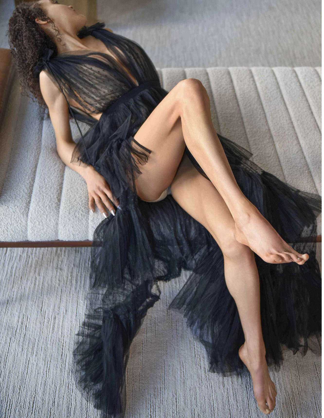A slim mixed woman wearing a black, sheer, and ruffled nightgown, with her legs crossed and her left hand resting on her hip.