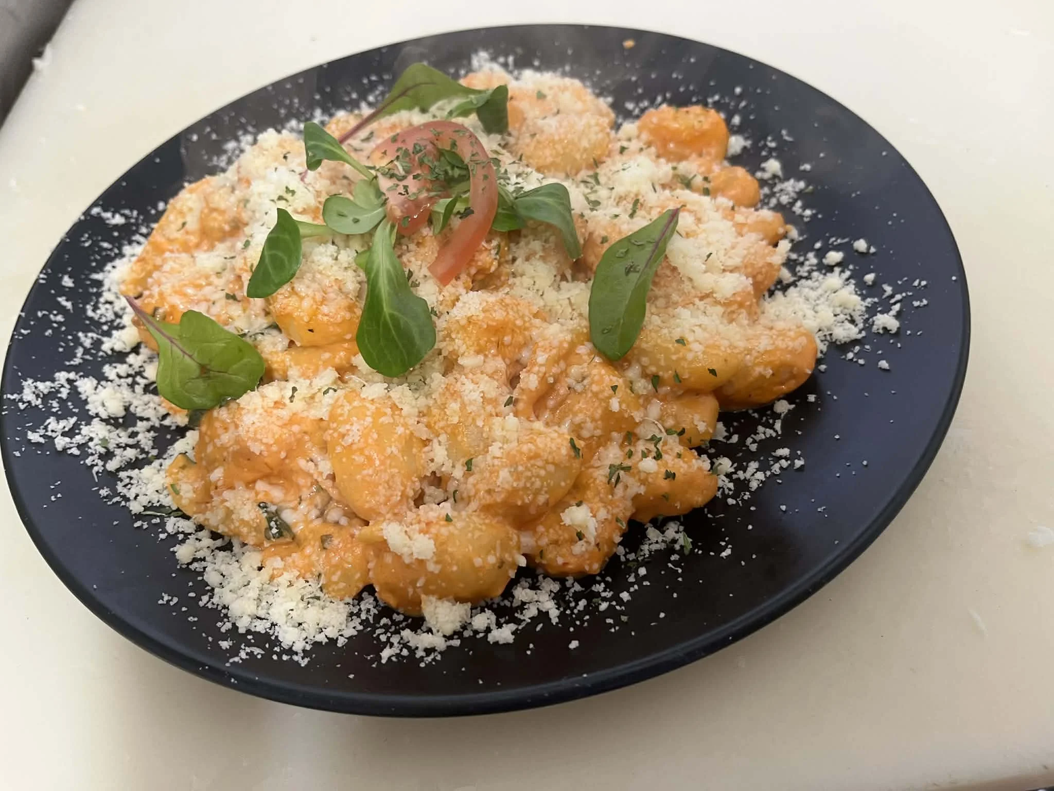 Plate of gnocchi pasta with tomato sauce, grated cheese, basil, and cherry tomato garnish on black plate.