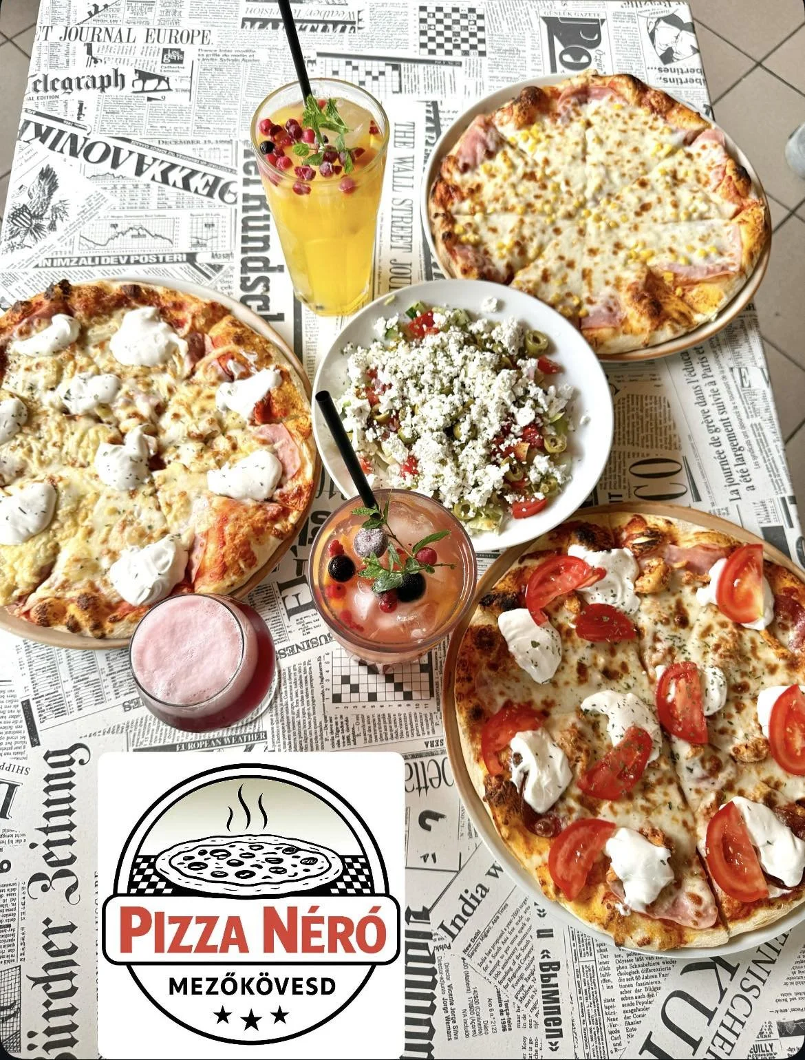 Table set with four pizzas topped with different ingredients, two colorful cocktails with fruit garnishes, and a side salad with feta cheese and vegetables. The table has a newspaper-themed tablecloth with a "Pizza Néro" logo in the foreground.