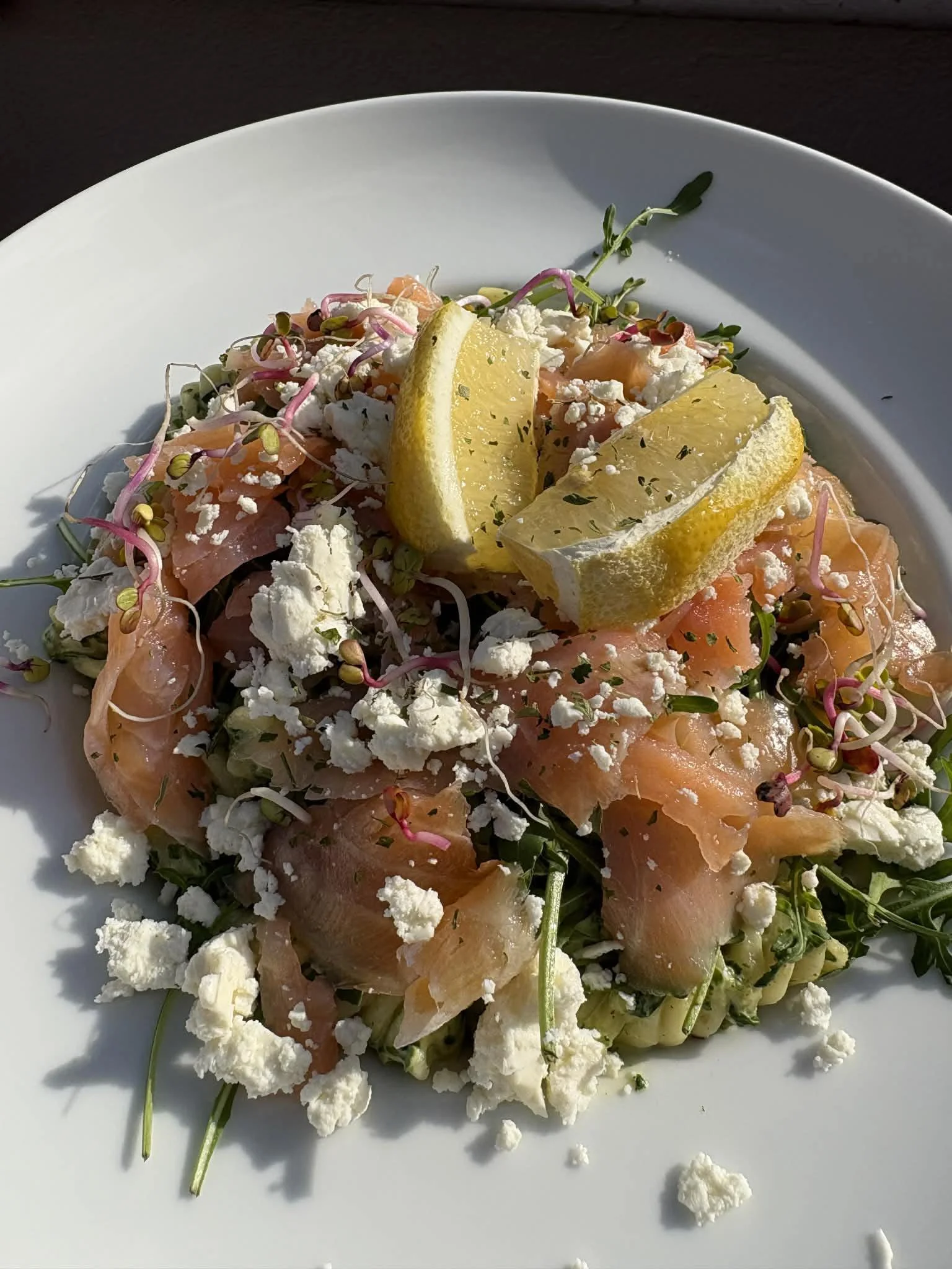 Salmon sashimi salad topped with lemon wedges, crumbled cheese, microgreens, and herbs on a white plate.