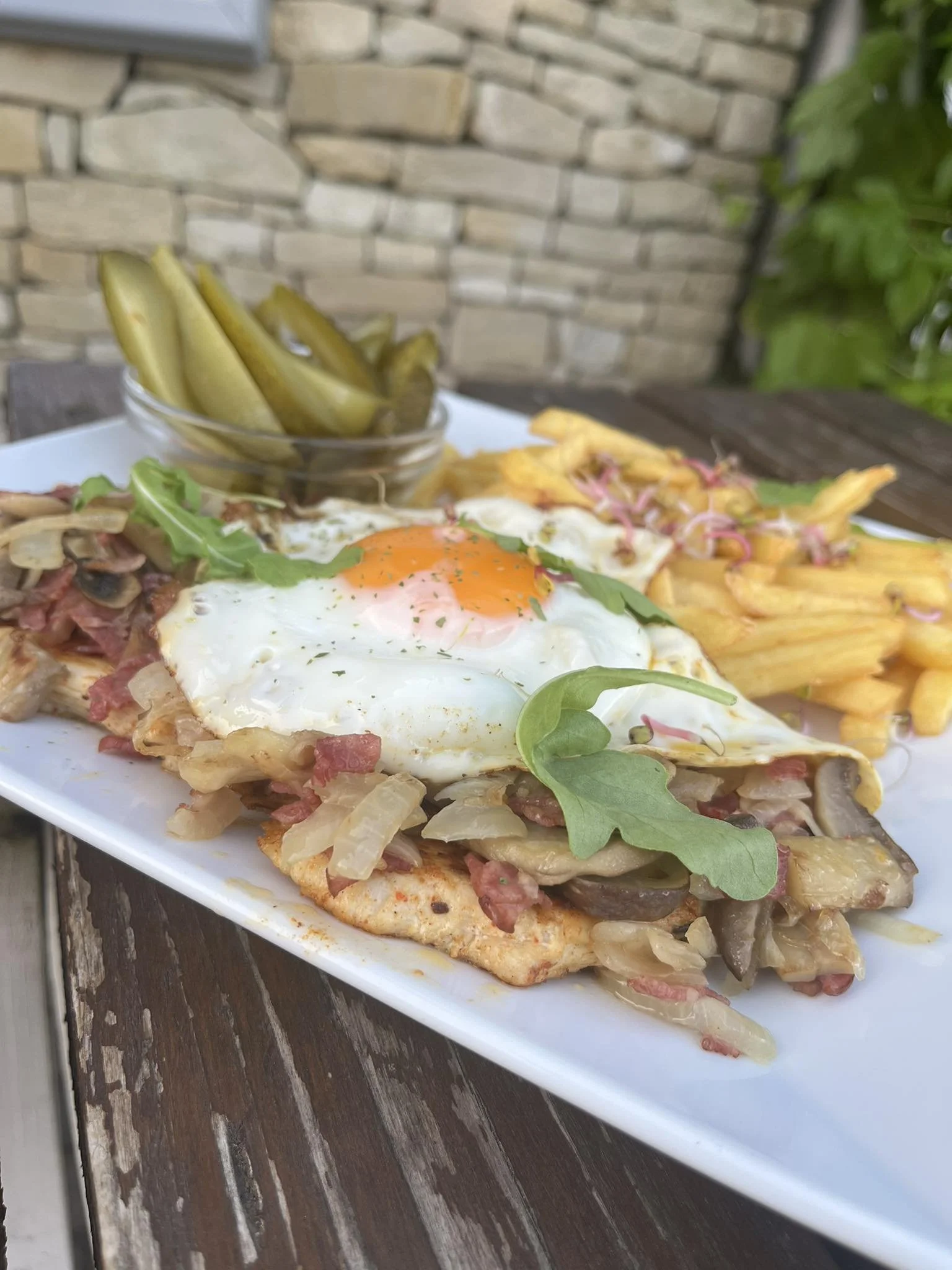 A plate of food with a fried egg on top of a bed of sautéed vegetables and meats, served with French fries in the background and pickles in a small glass bowl.