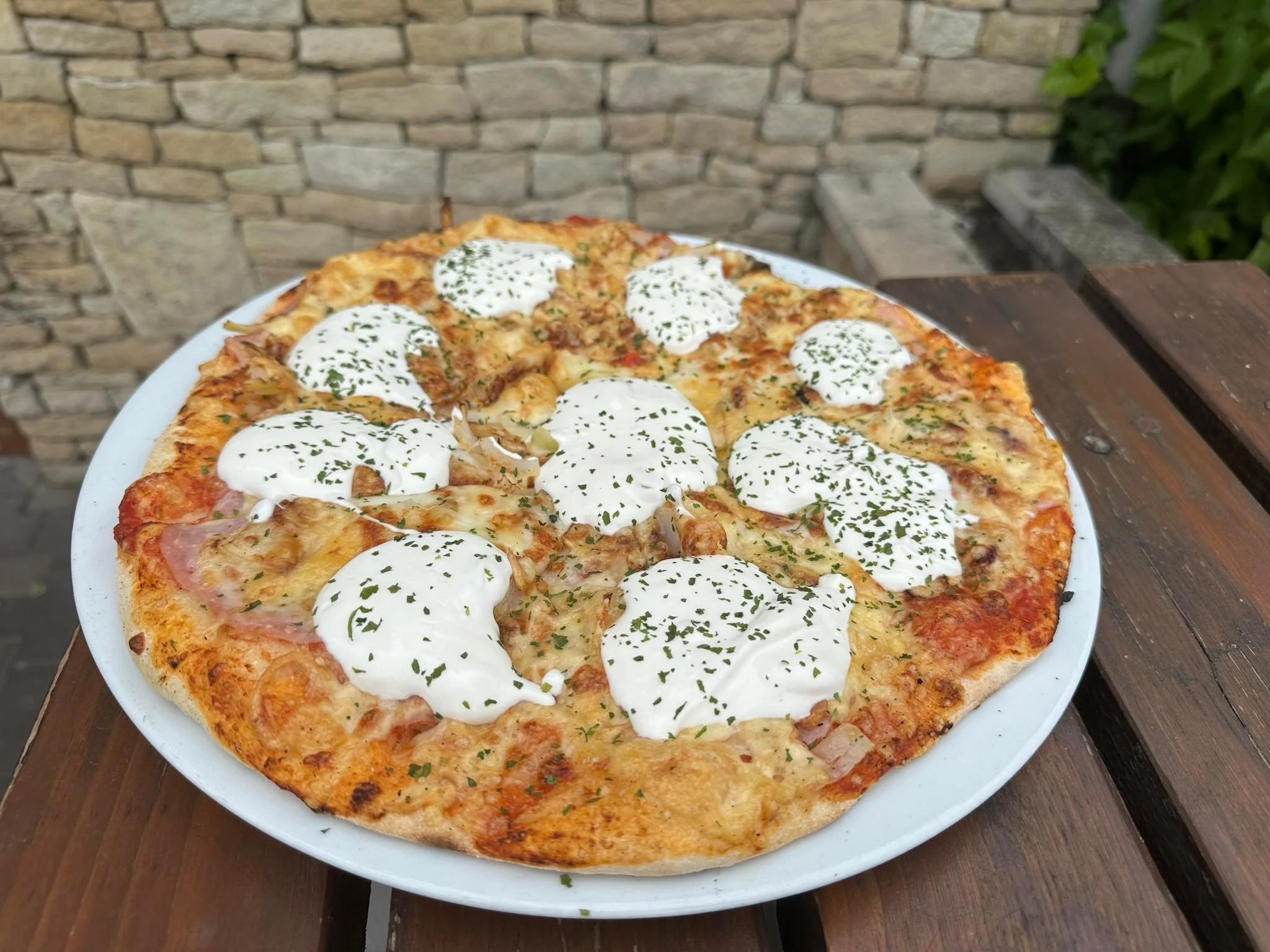 Pizza with a thin crust, topped with white sauce, herbs, and various toppings including chicken, bacon, and ham, served on a white plate on a wooden table outdoors.