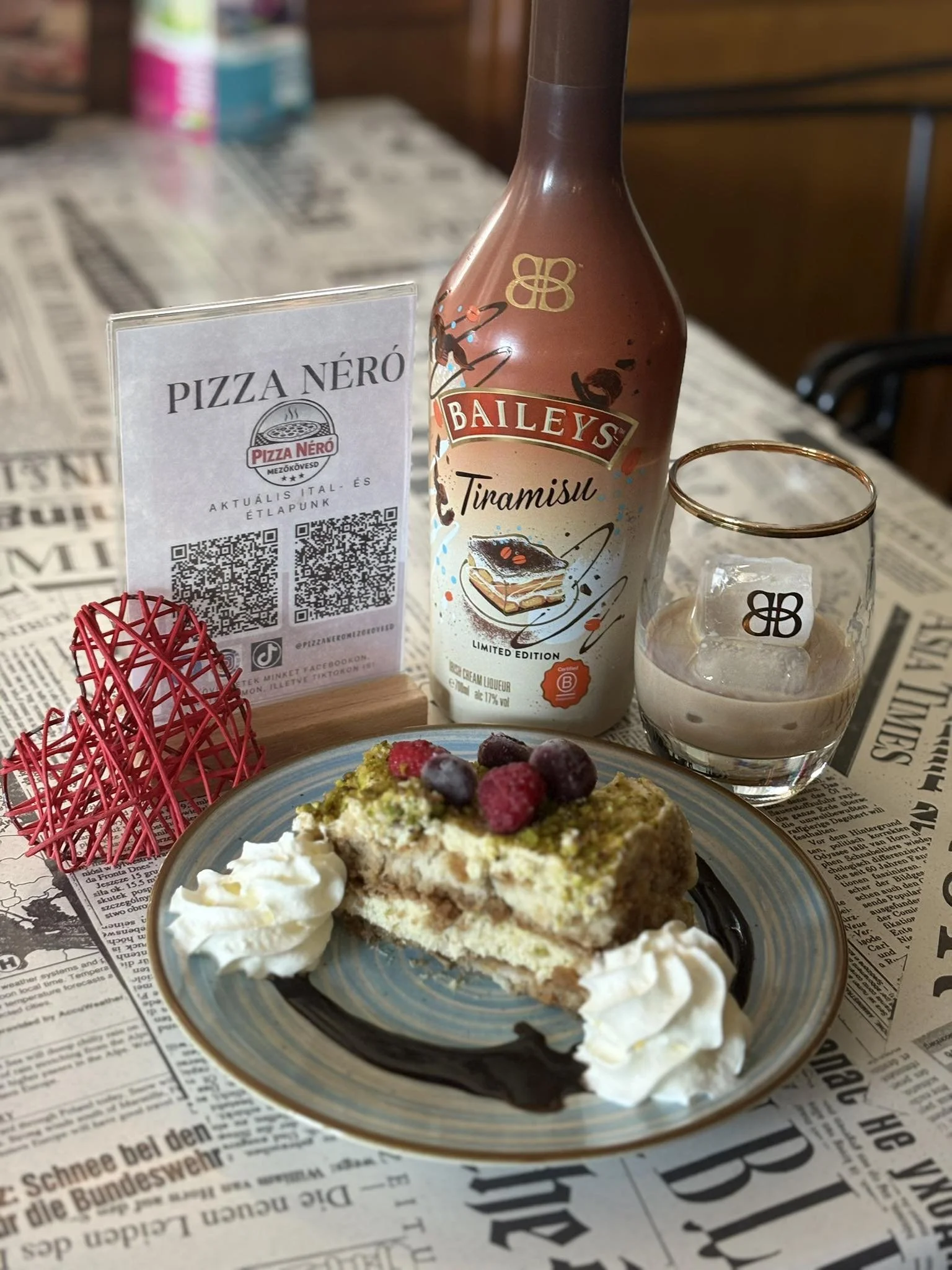A slice of tiramisu cake with berries and whipped cream on a blue plate, a glass of Baileys Irish Cream with ice, a red decorative heart, and a sign for Pizza Néro restaurant on a table covered with a newspaper-style tablecloth.