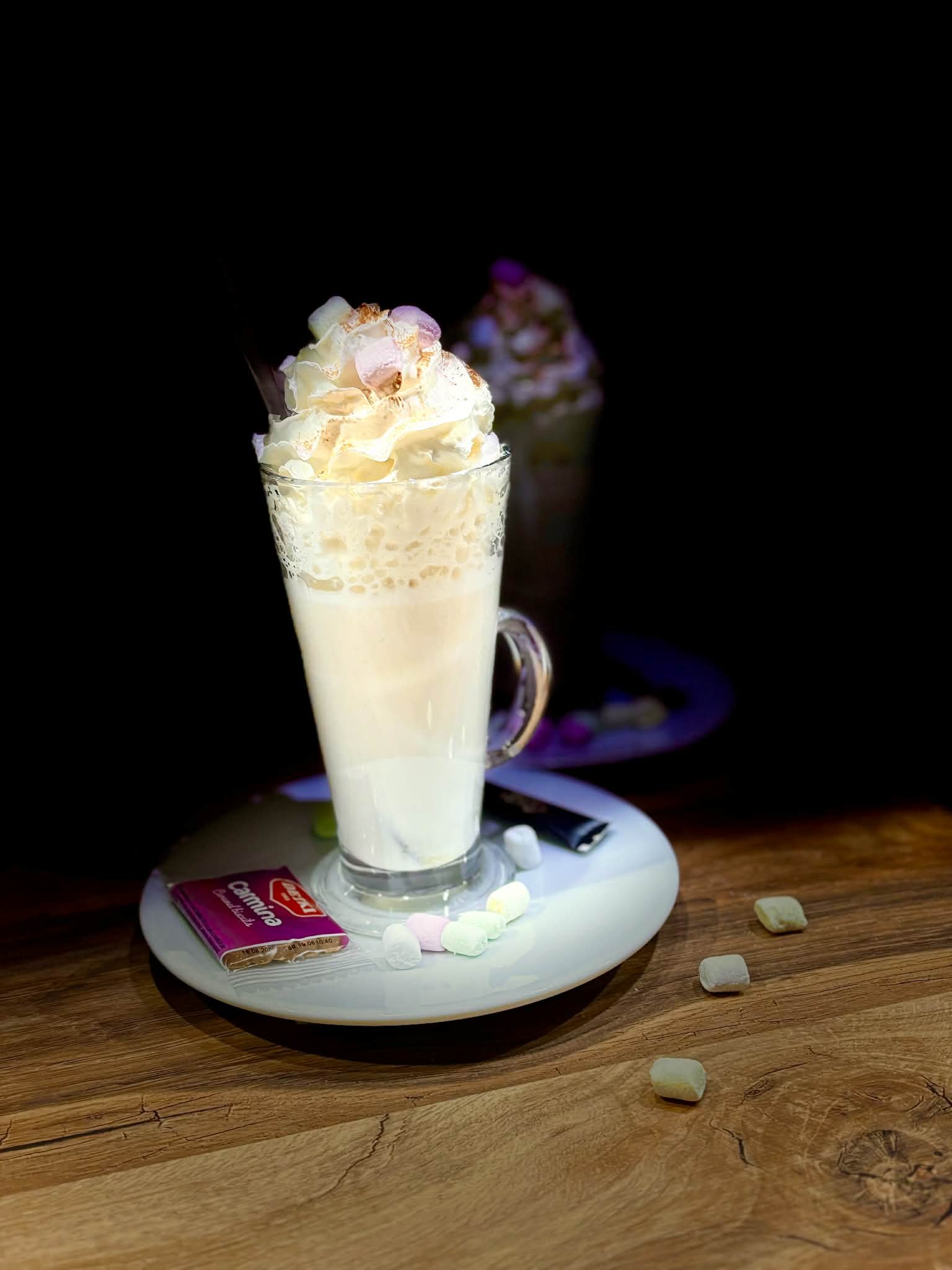 A tall glass of milkshake topped with whipped cream and marshmallows, placed on a white plate with a packet of caramel candies and marshmallows scattered around, on a wooden table with a dark background.