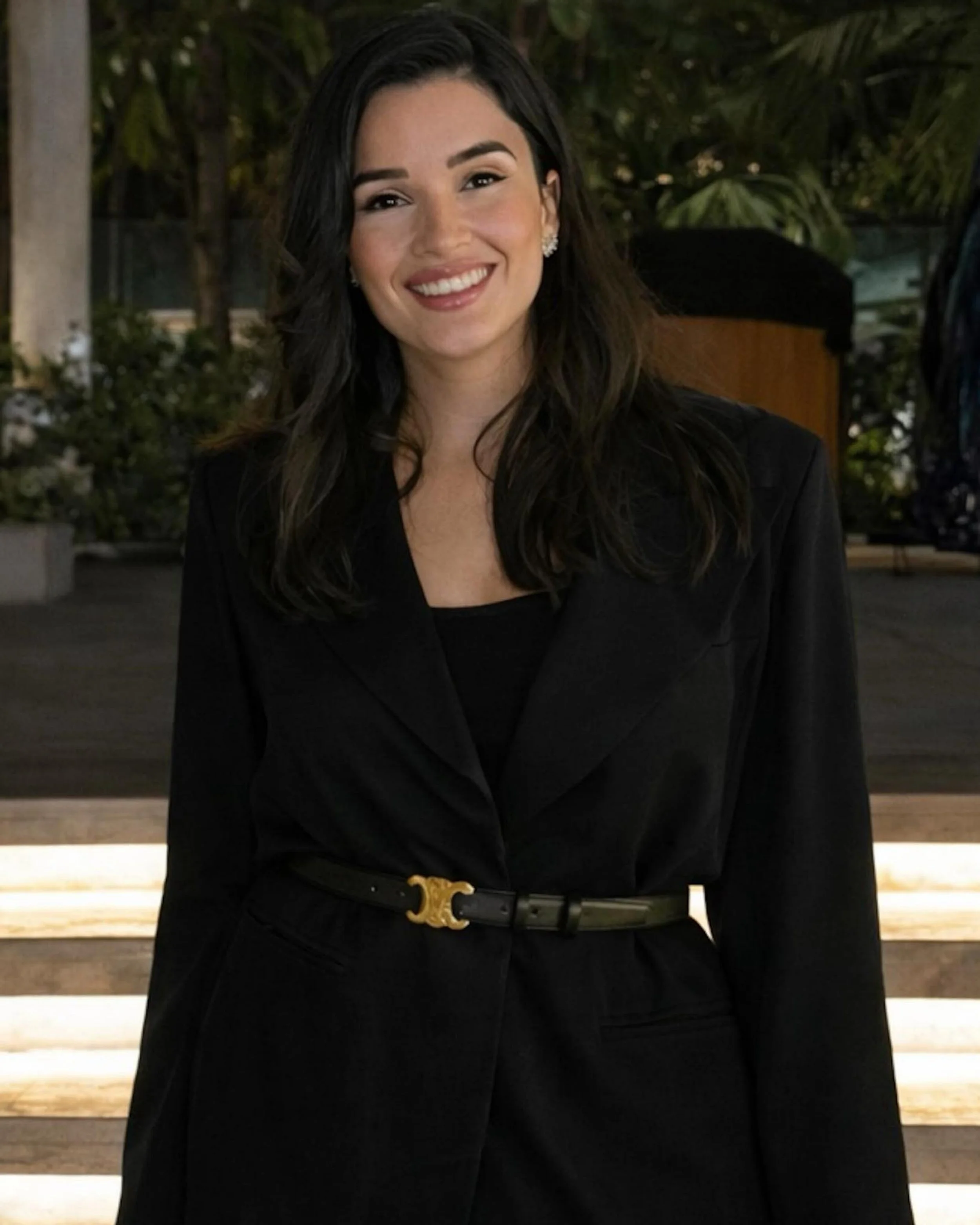 A smiling young woman with dark hair wearing a black blazer and a belt with a gold buckle, posing outdoors at night.