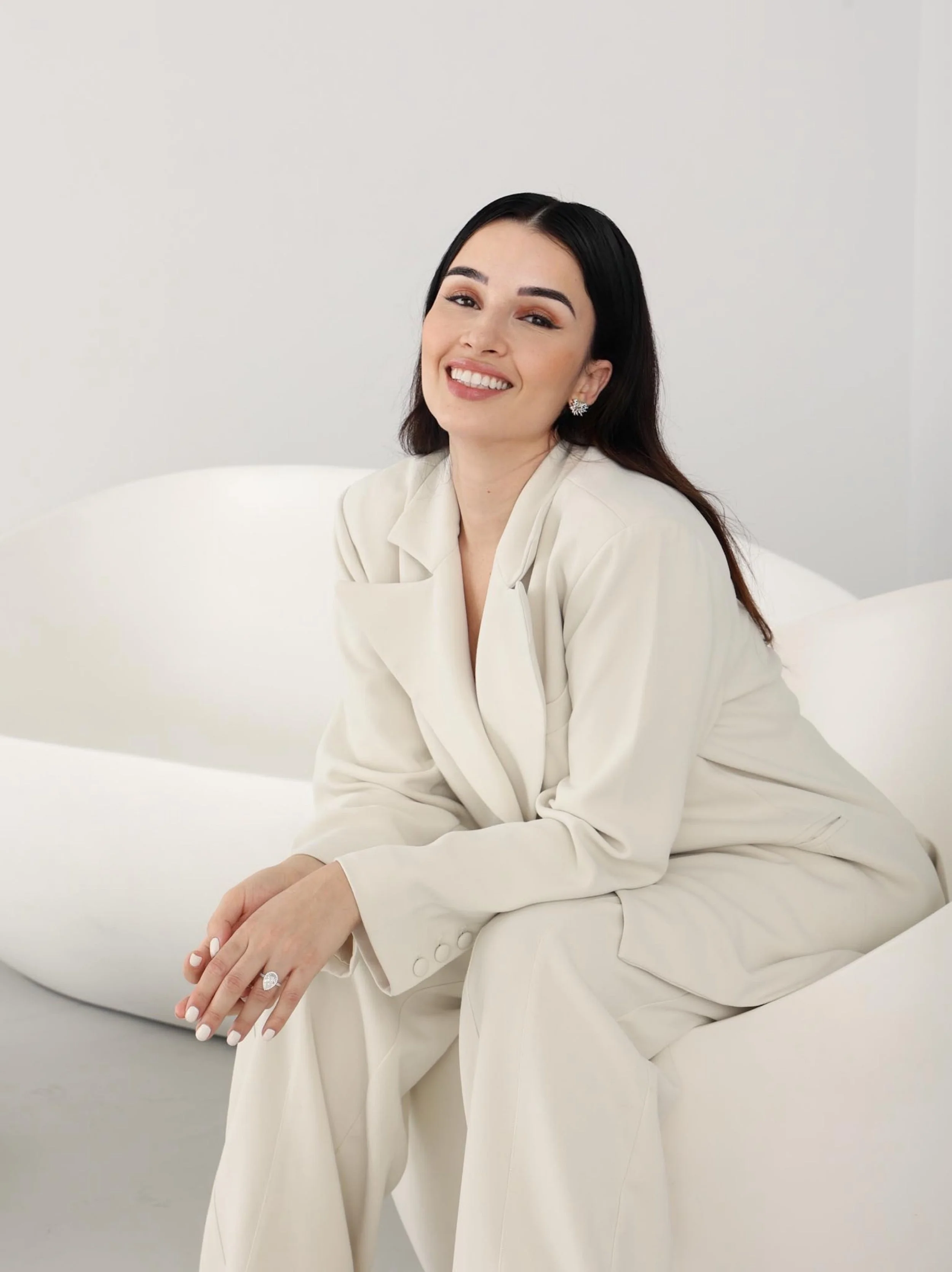 Young woman with dark hair and earrings sitting on a white sofa, smiling in a minimalistic white room.