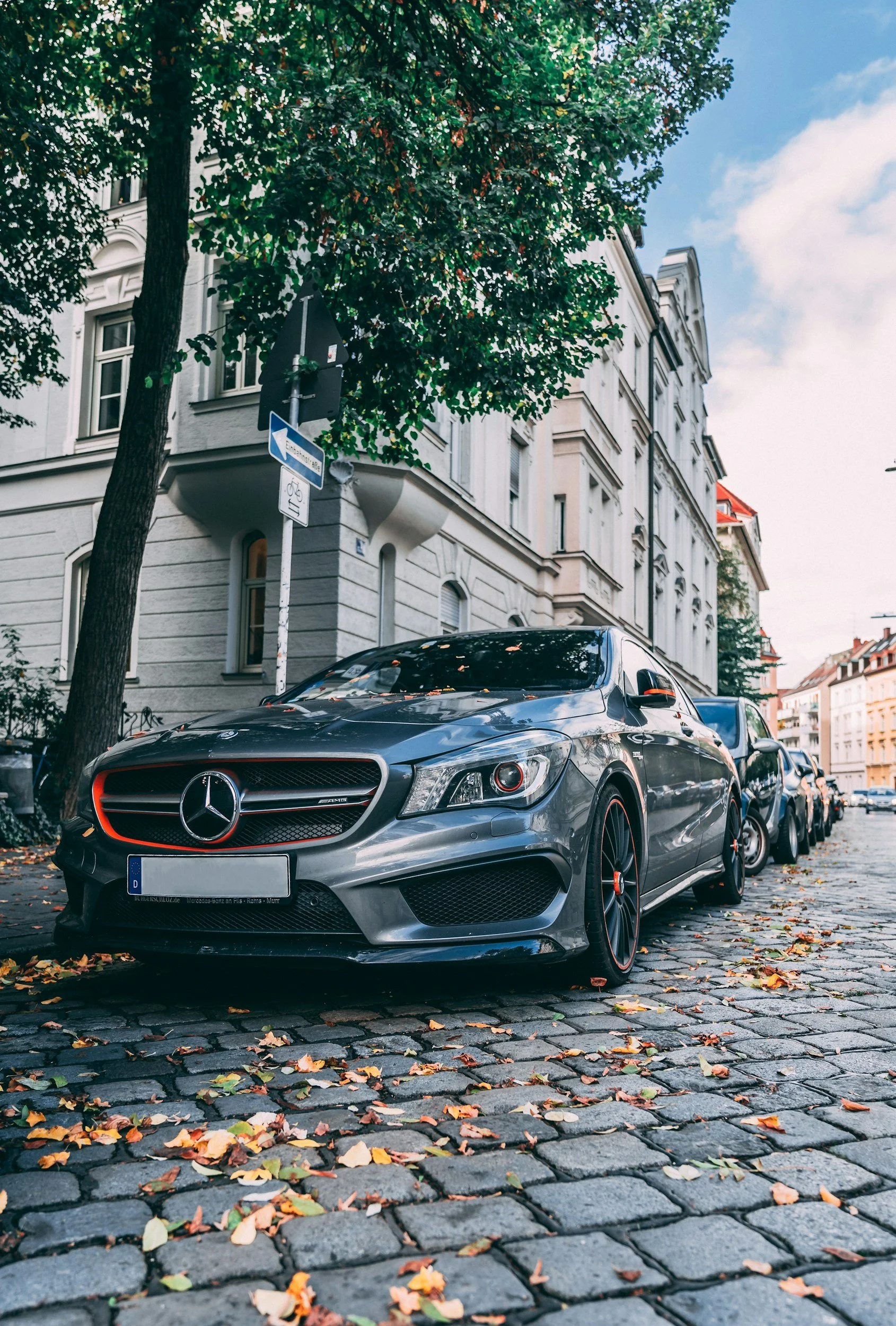A silver Mercedes-Benz parked on a cobblestone street, with autumn leaves scattered on the ground and buildings with classic architecture in the background.