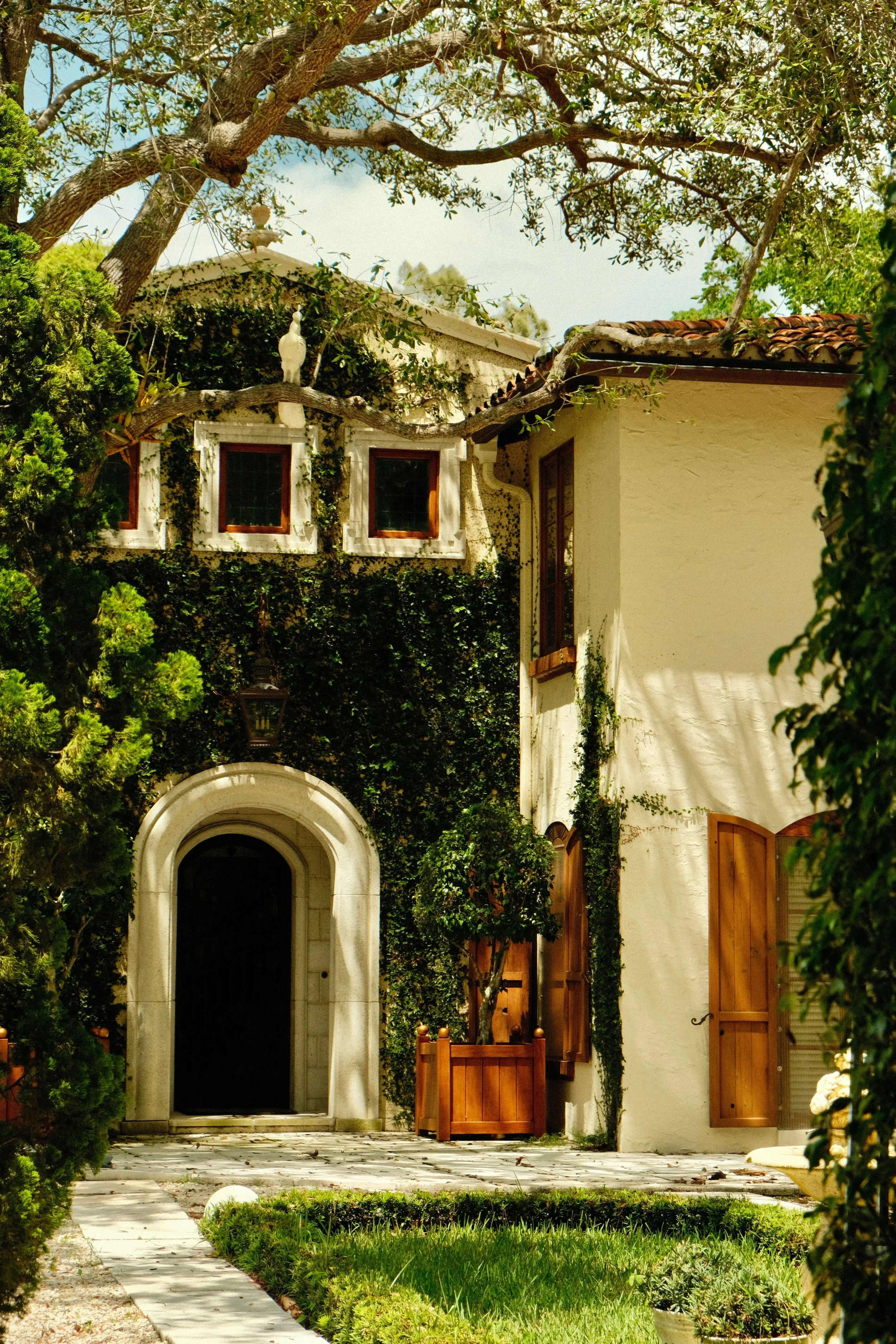 A house covered in green ivy with wooden window shutters, a stone archway entrance, and a tree with a white bird sculpture on its branch in the front yard.